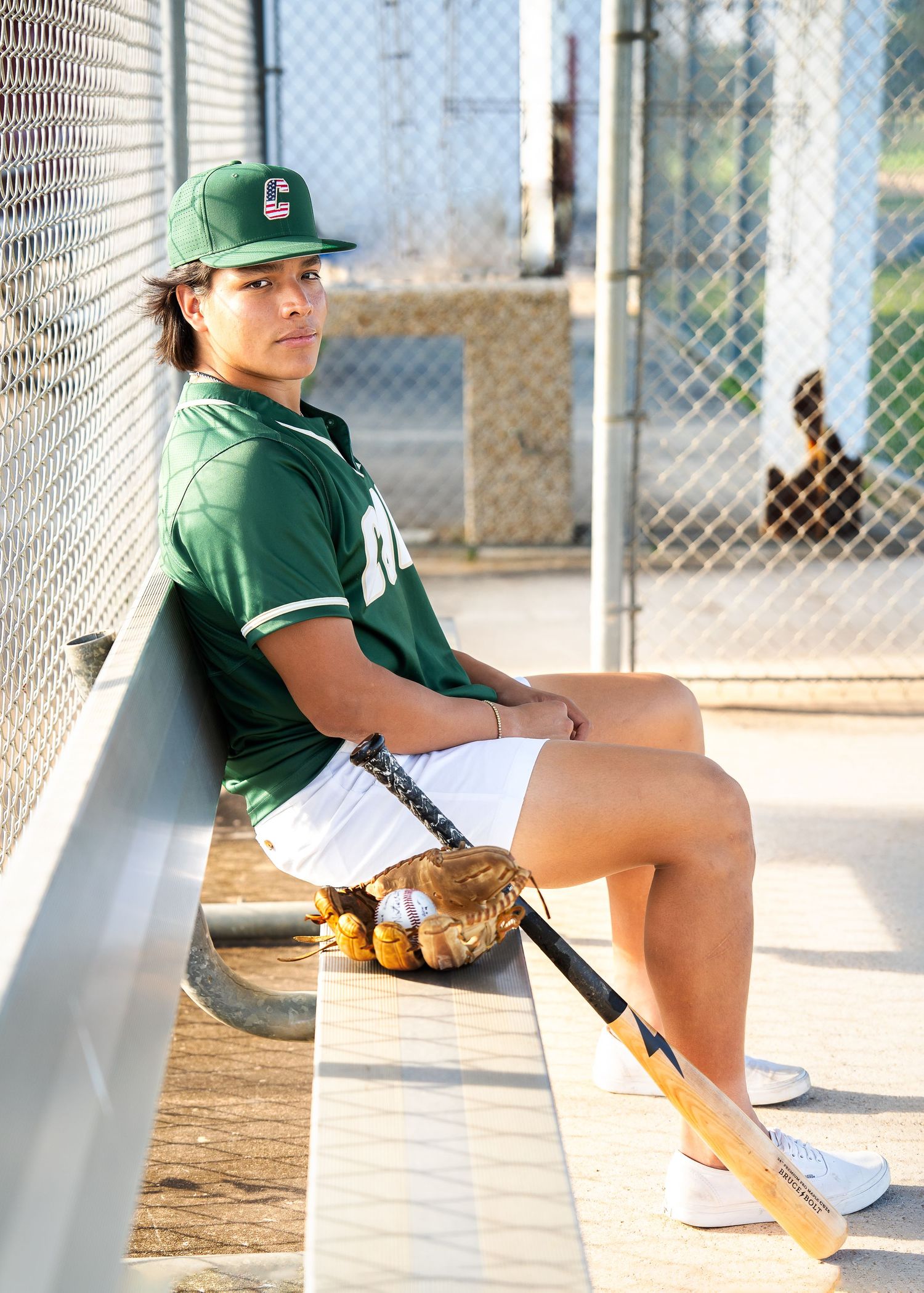 high school senior boy sitting in baseball dugout with baseball bat and mitt next to him