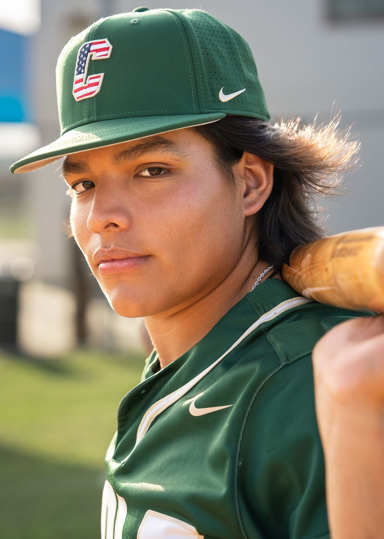 high school senior boy wearing green baseball jersey and baseball hat holding a baseball bat behind his neck