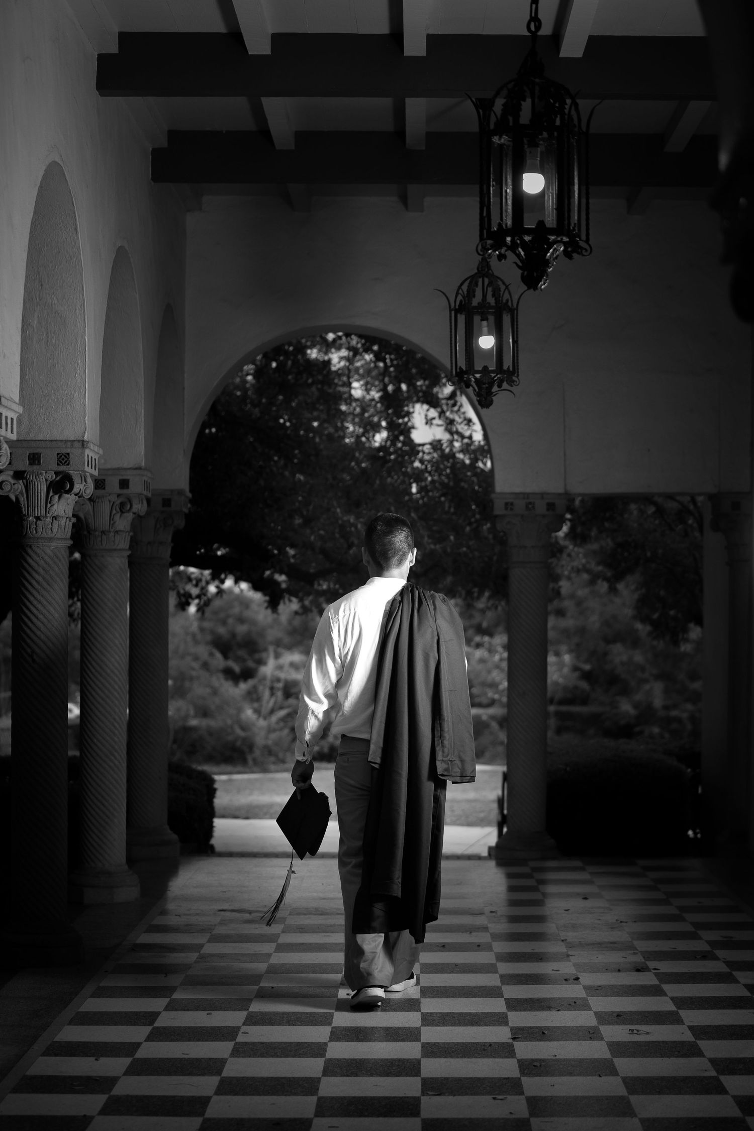 black and white portrait of high school senior boy walking away across the porch at Landa Library with cap and gown