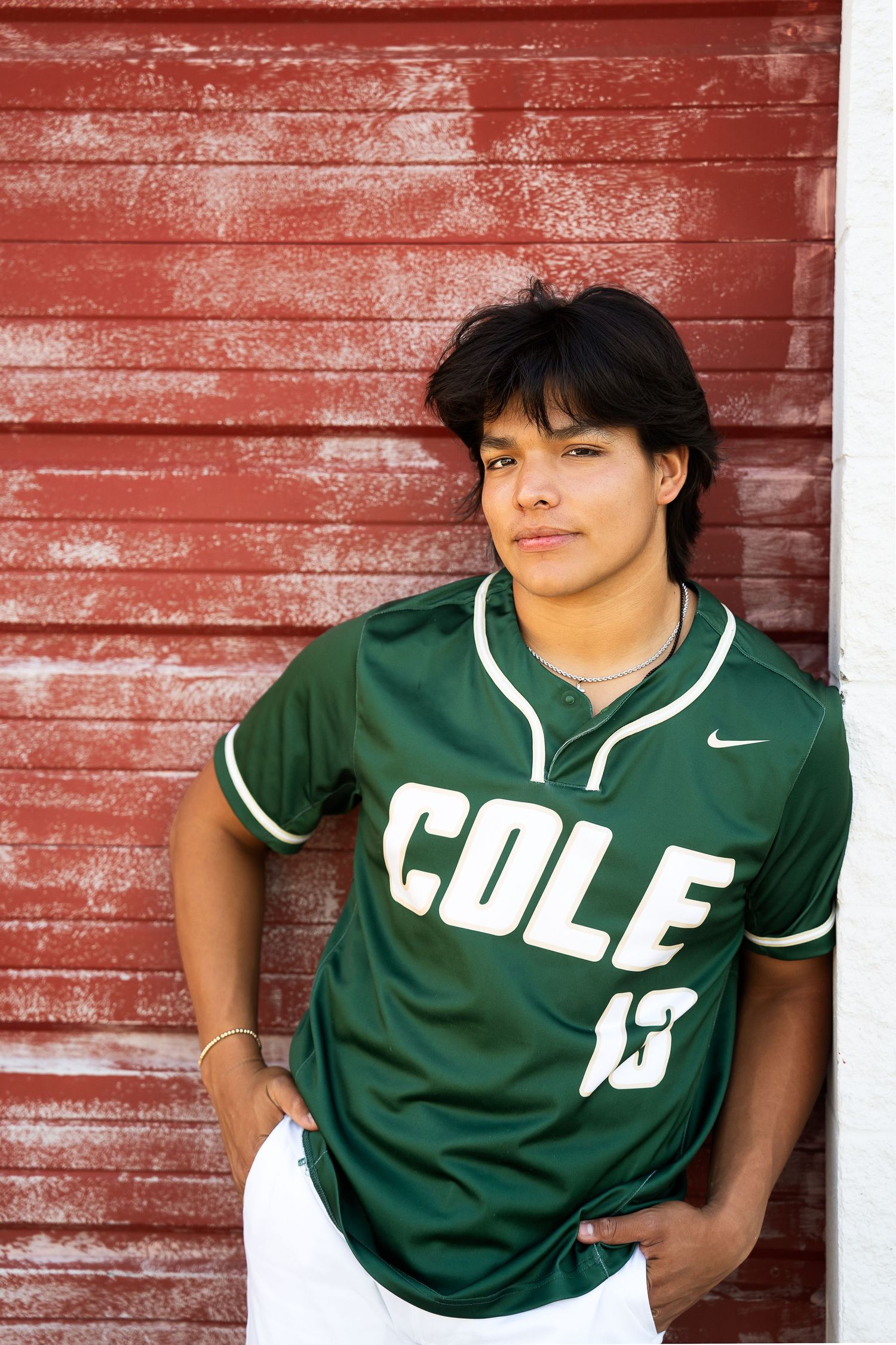 high school senior boy wearing green baseball jersey standing against a red wall in San Antonio Texas