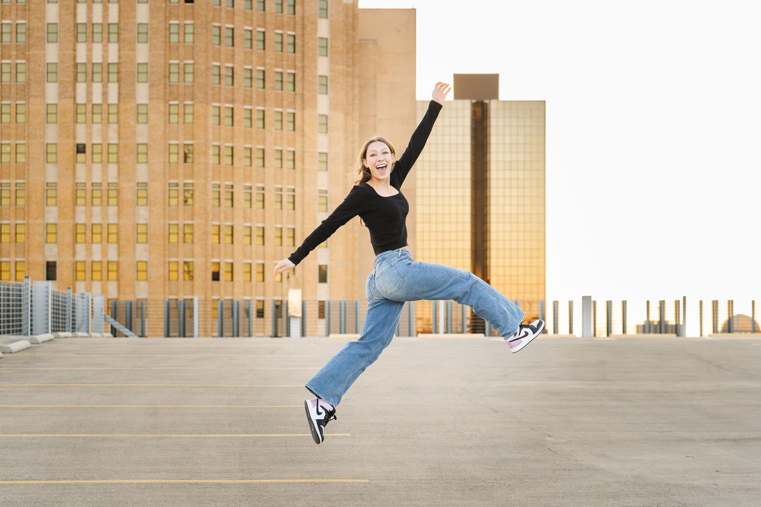 high school senior girl in jeans and black sweater leaping in the air on roof of parking garage with city behind
