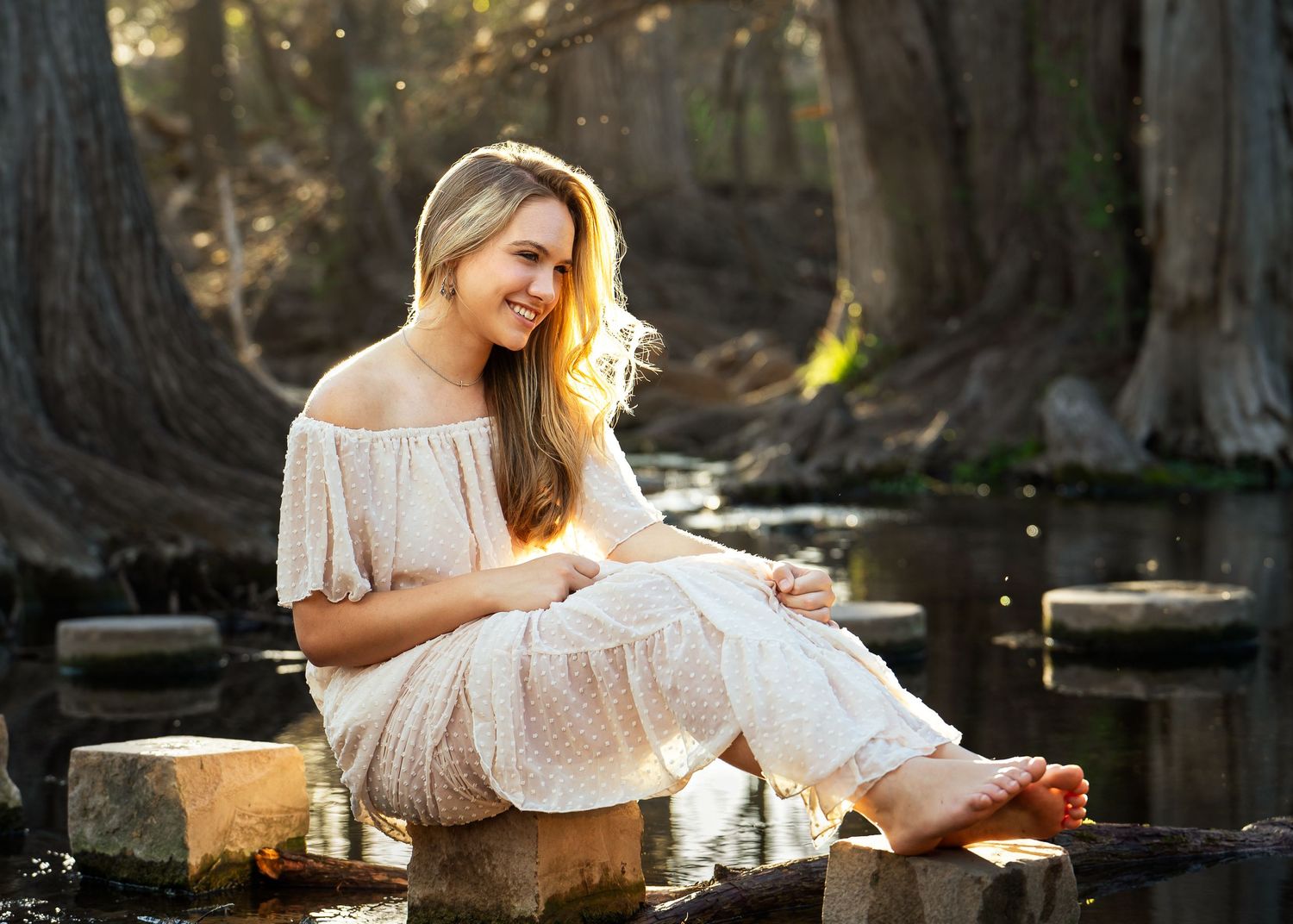 high school senior girl in cream flowy dress sitting on rocks near Cibolo Creek in Boerne Texas