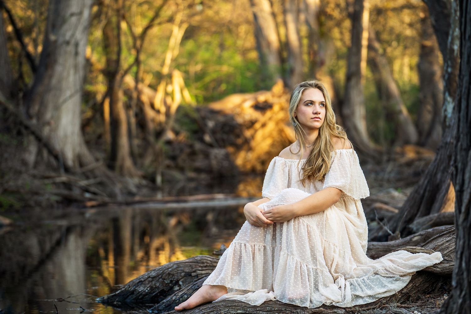 high school senior girl in cream long dress sitting near Cibolo Creek at Cibolo Creek Nature Center