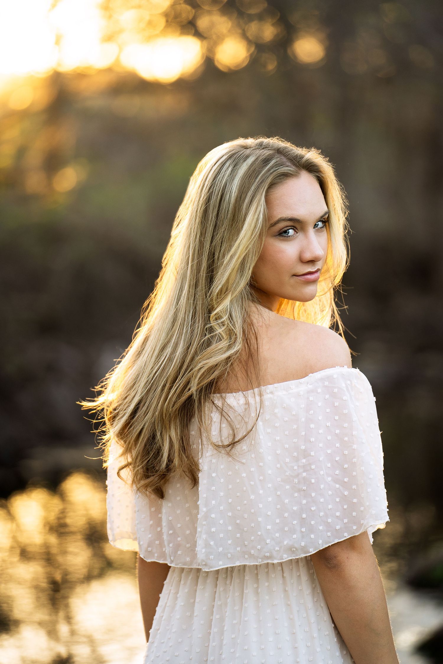 high school senior girl wearing an off the shoulder cream dress looking over her shoulder with golden sunlight in Boerne