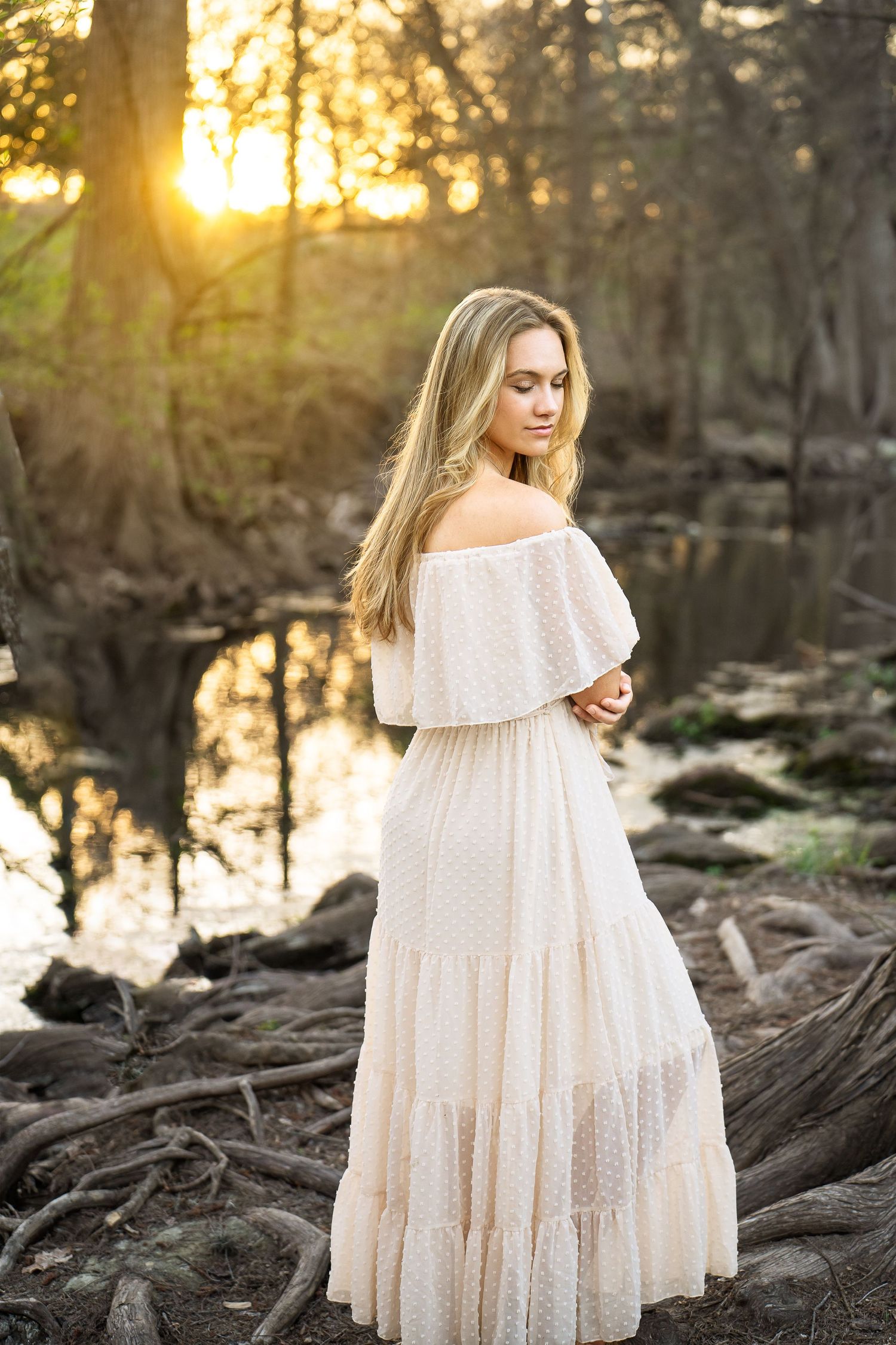 high school senior girl wearing white off the shoulder dress standing in front of golden sunset at Cibolo Creek