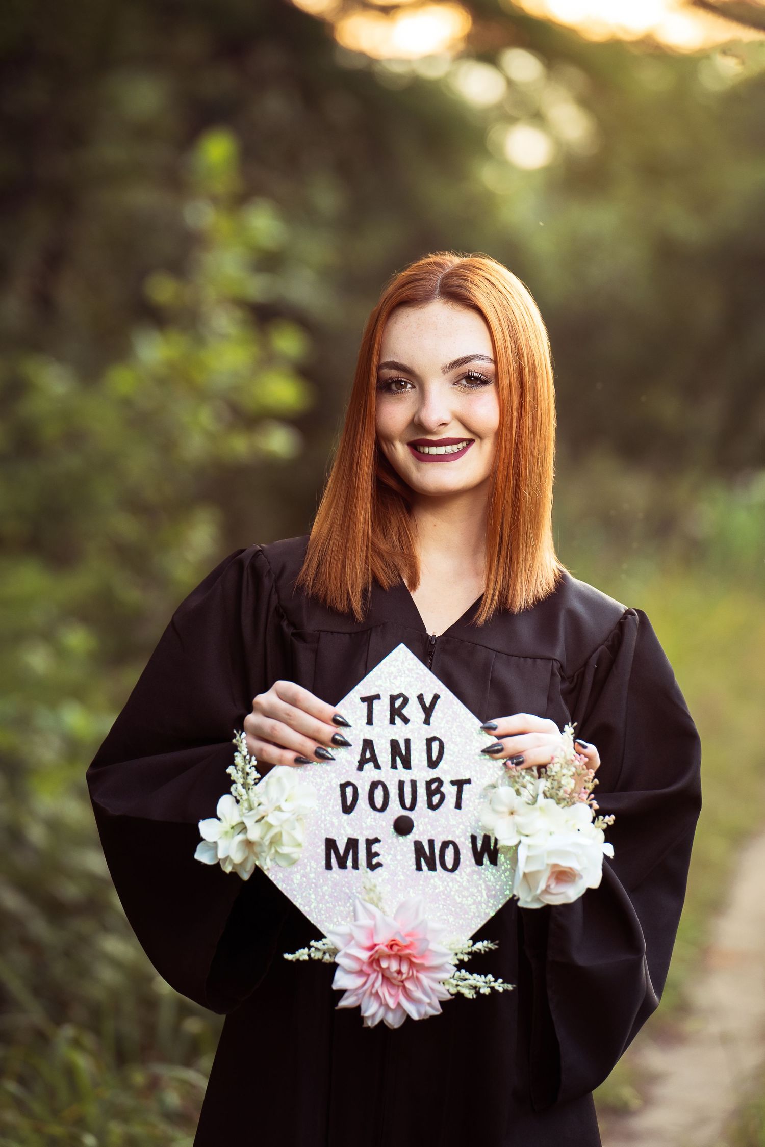 high school senior girl wearing black graduation gown holding cap with flowers and saying on walking path