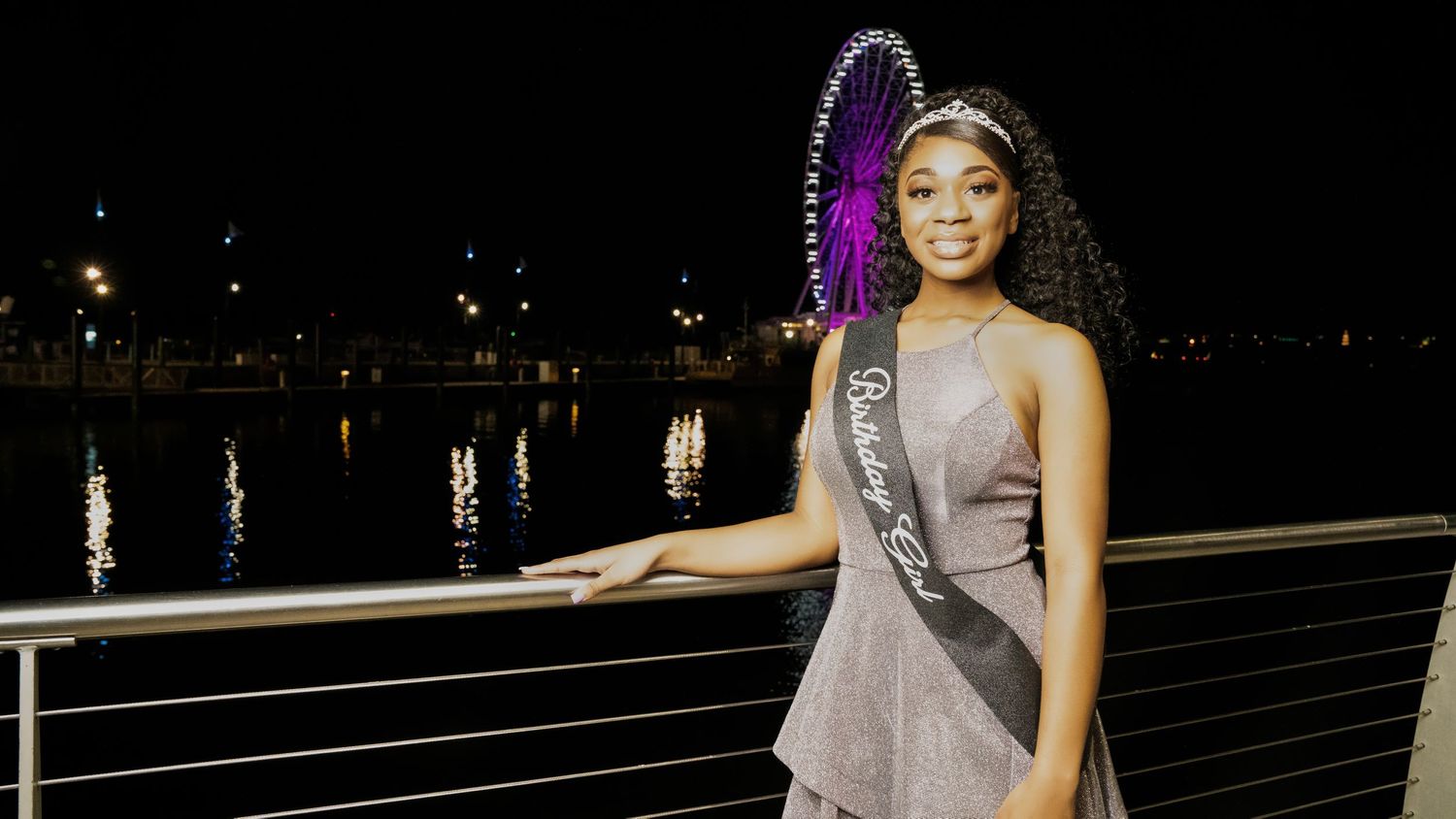 Young woman in silver dress wearing queen sash posing by waterfront at night.
