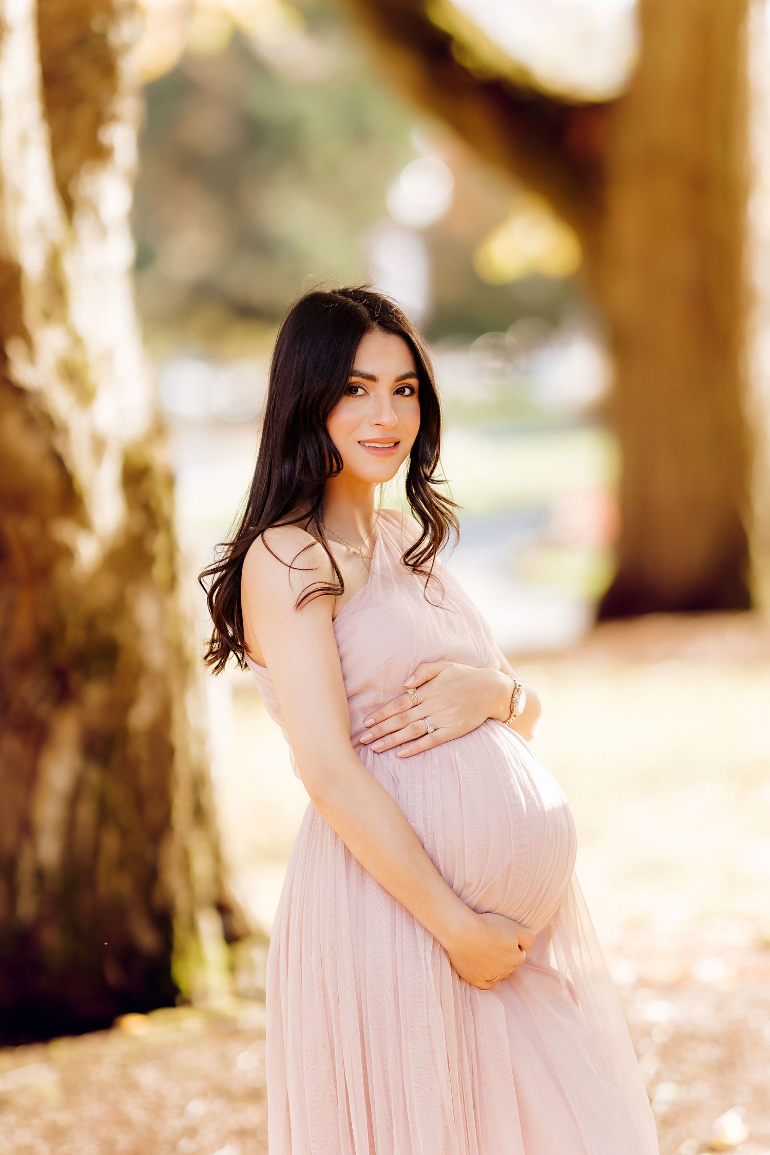 Woman in pink dress cradling baby bump in sunlit outdoor setting.