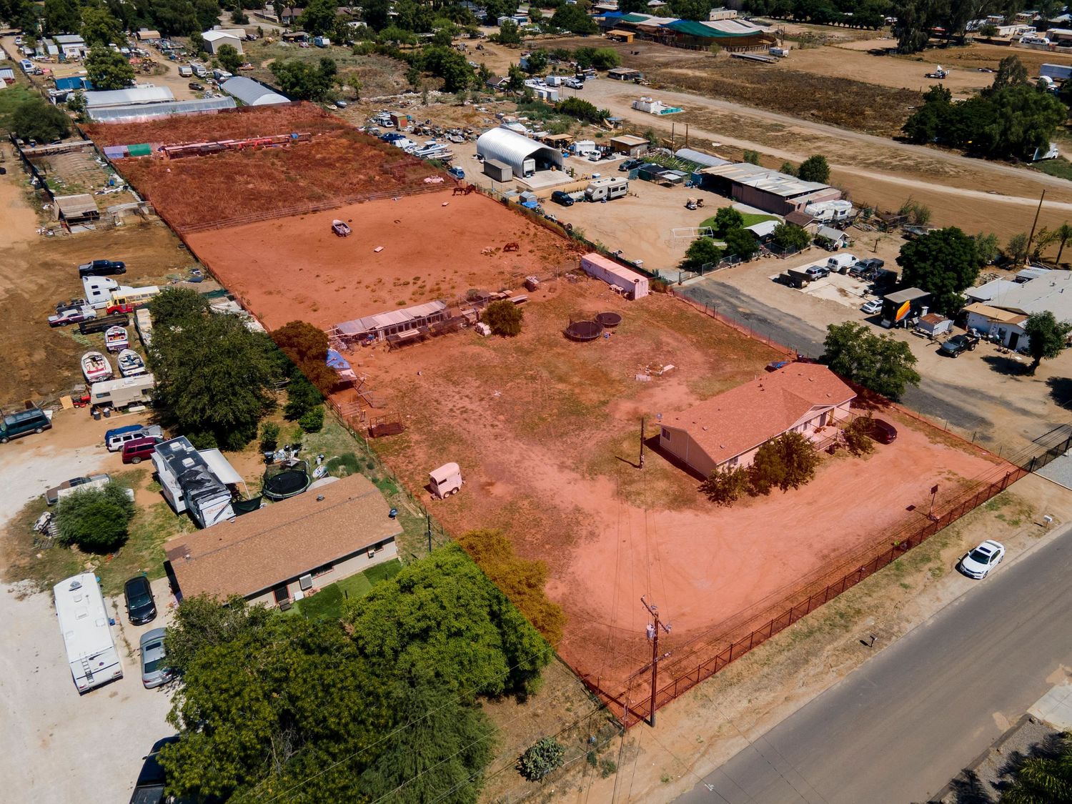 Aerial drone view of a large red dirt lot with structures and surrounding rural neighborhood.