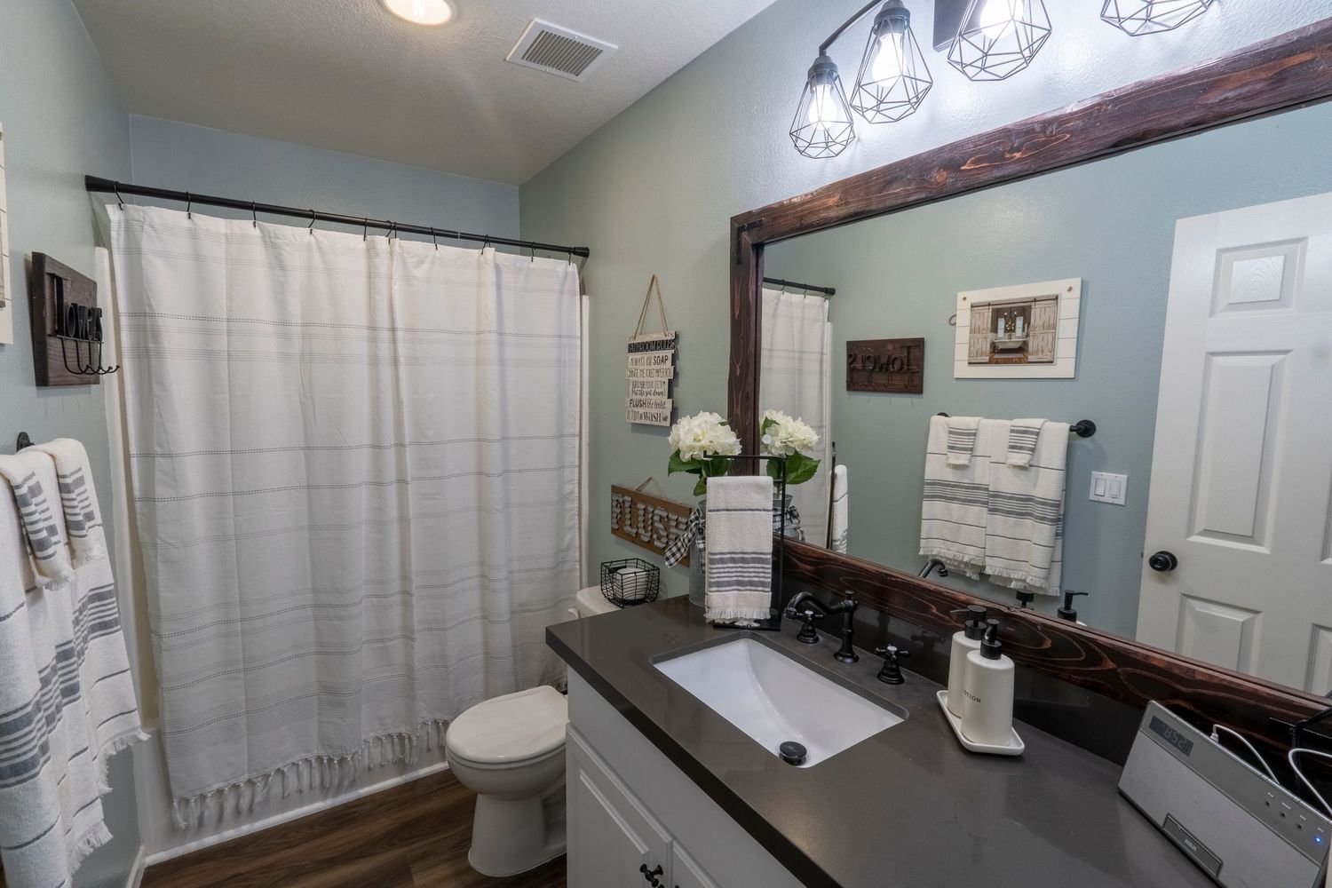 Modern farmhouse bathroom with dark wood-framed mirror, white shower curtain, and stylish pendant lighting.