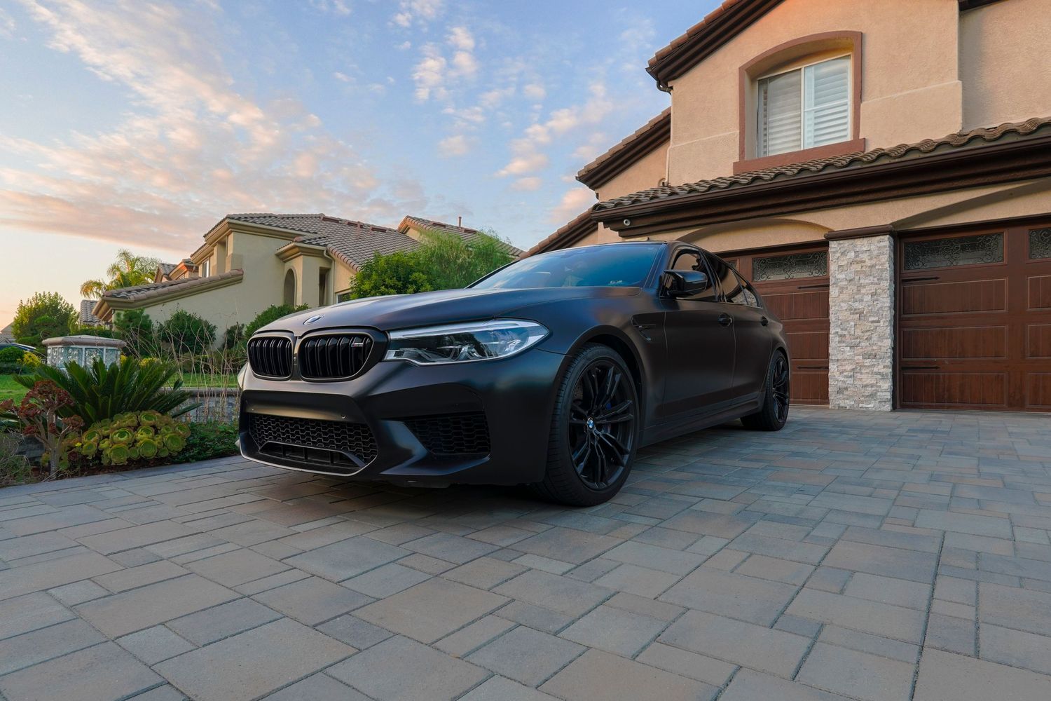 Dark matte BMW M5 parked on a paver driveway in front of a luxury home at sunset.
