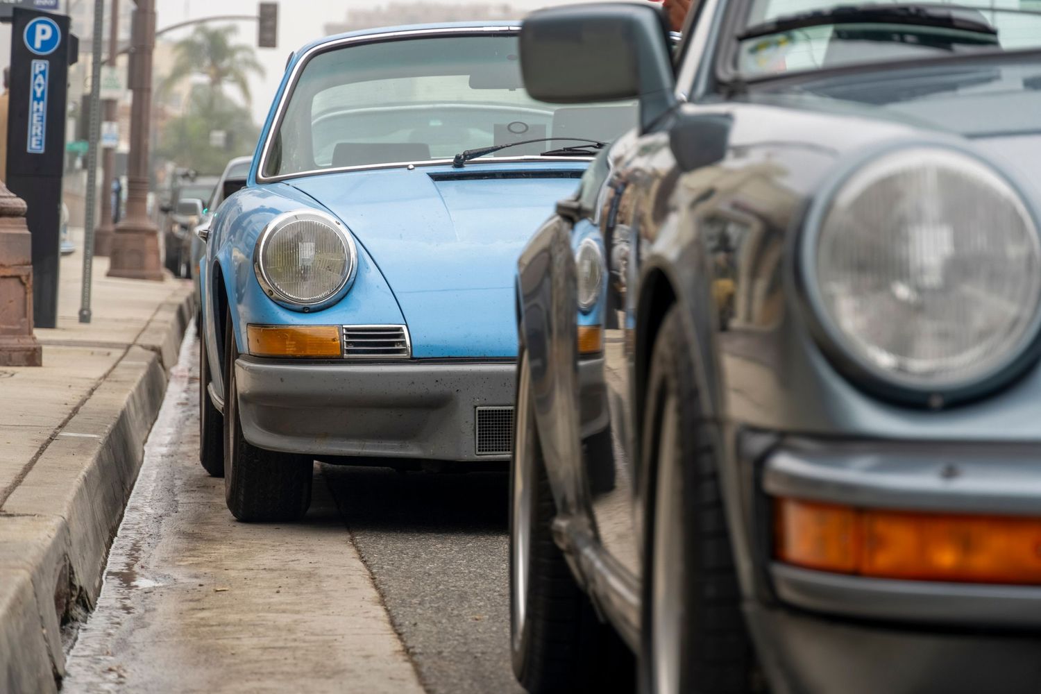 Two classic Porsche 911 sports cars parked on a city street, one in light blue and one in dark gray.