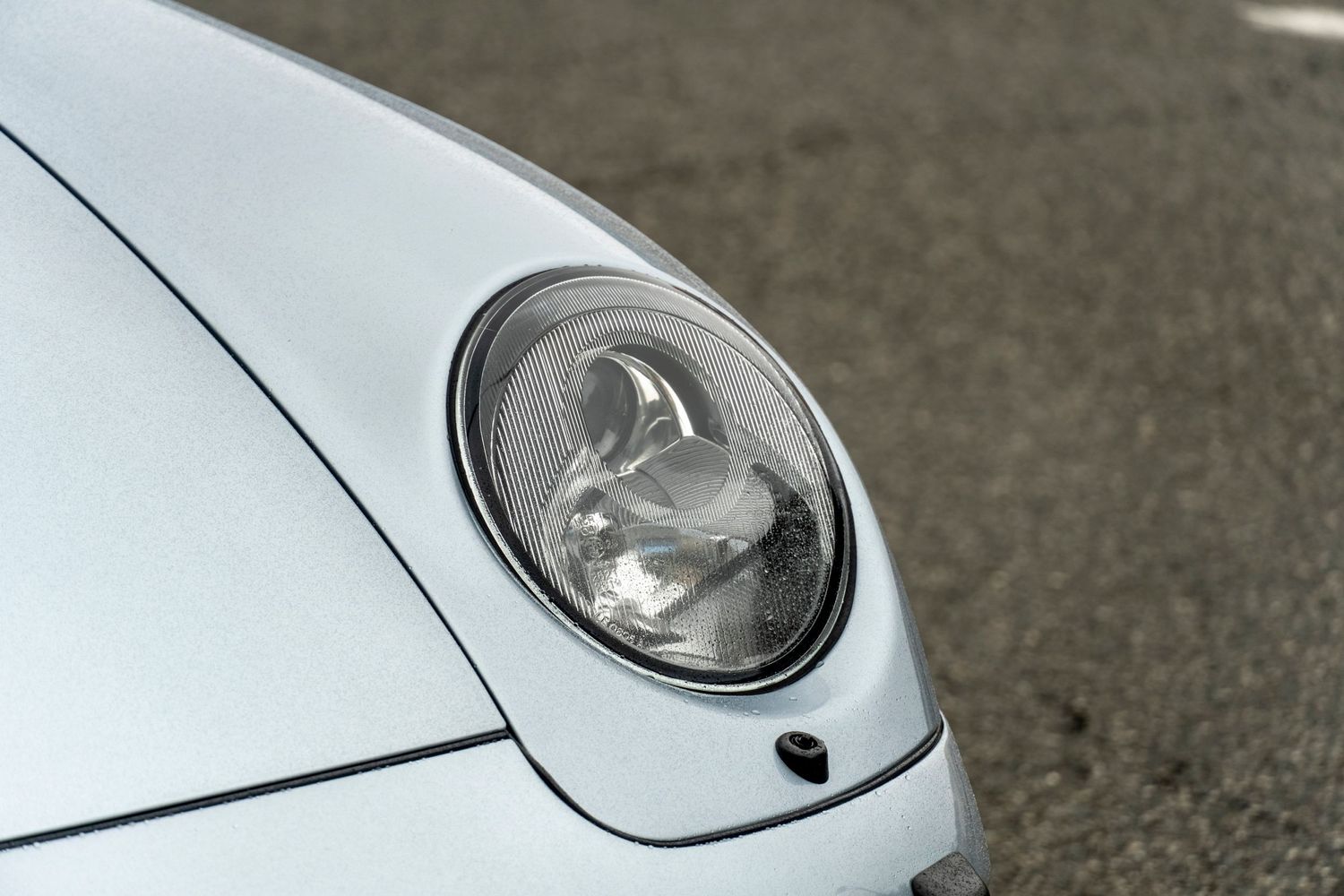 Close-up of a round headlight on a silver Porsche 911 parked on pavement.