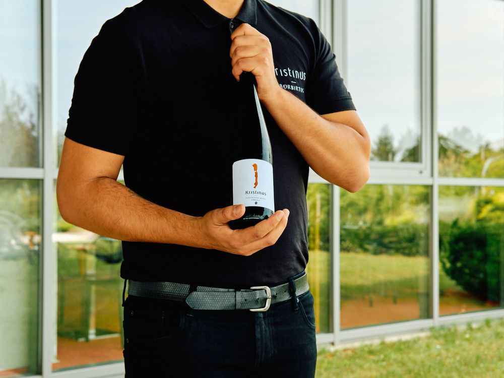 Server in black uniform holding a wine bottle against glass window background.