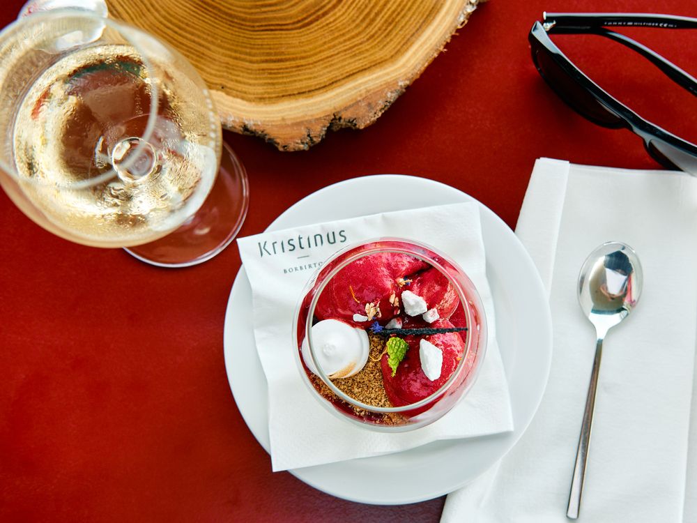 Table setting with wine glass, wooden coaster and red dessert on white plate.