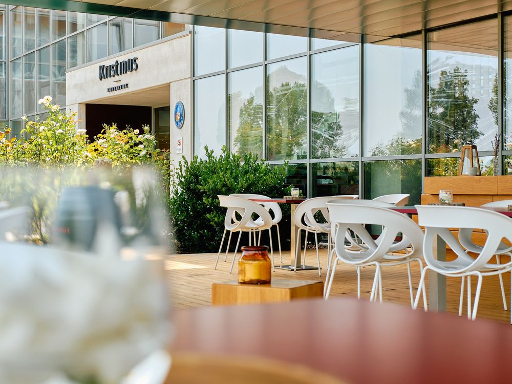 Modern restaurant interior with white chairs and large glass windows overlooking outdoor greenery.