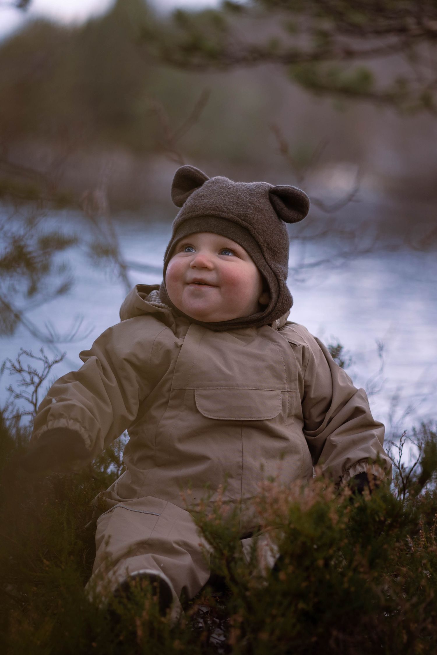 Small figure in tan snowsuit and bear hat smiles in winter nature setting.