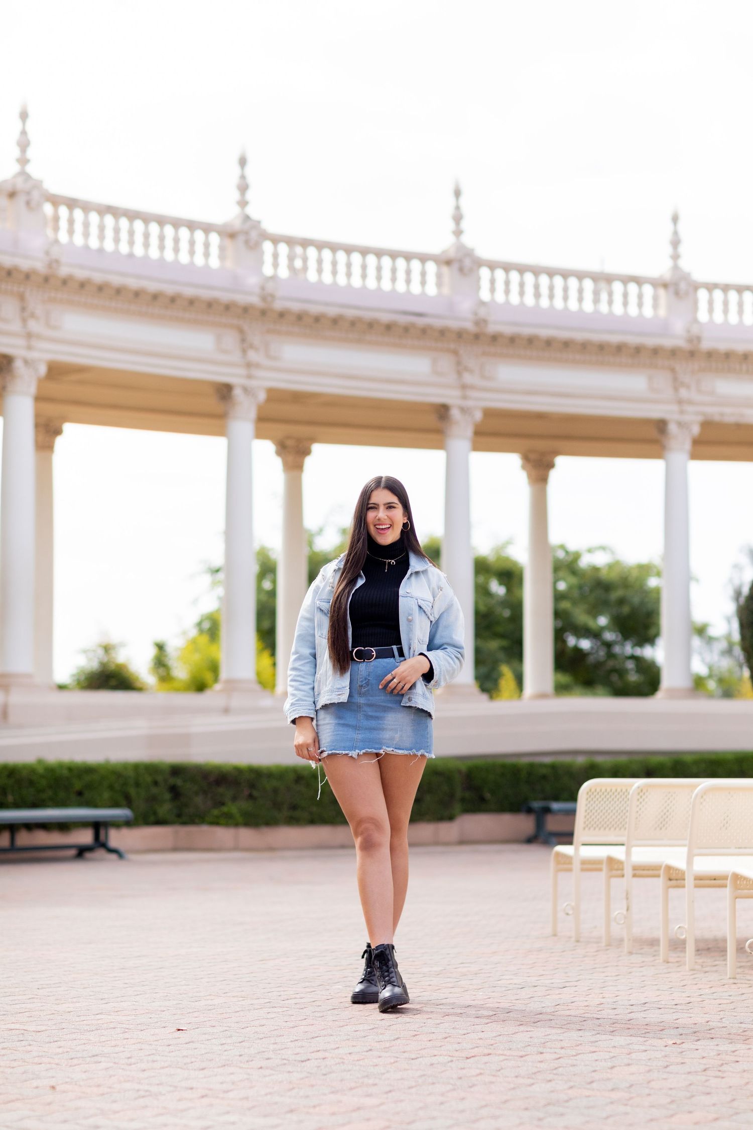 A high school senior in denim skirt and jean jacket posing under white columned architecture.