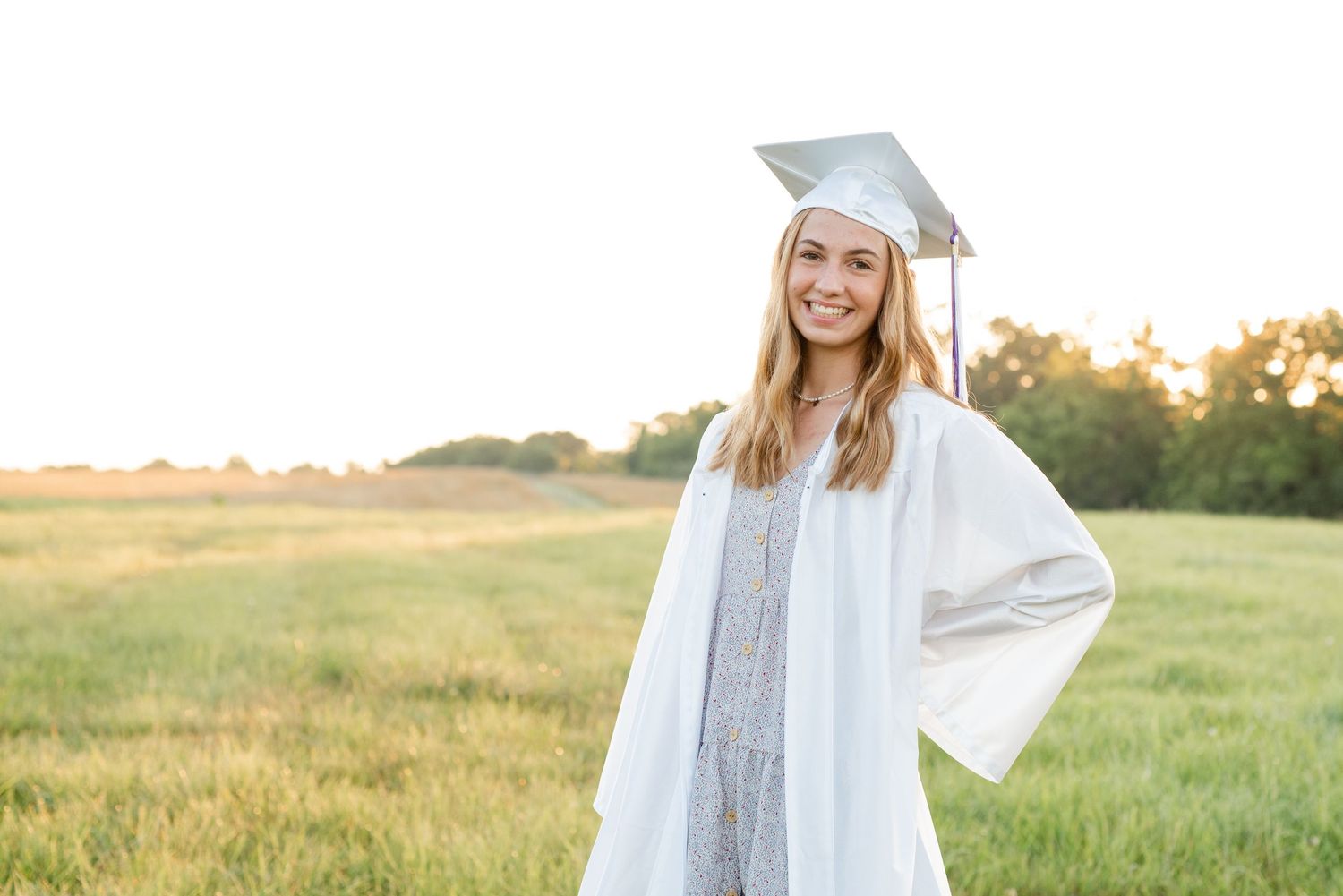 Graduate in white cap and gown poses in sunny field during golden hour.