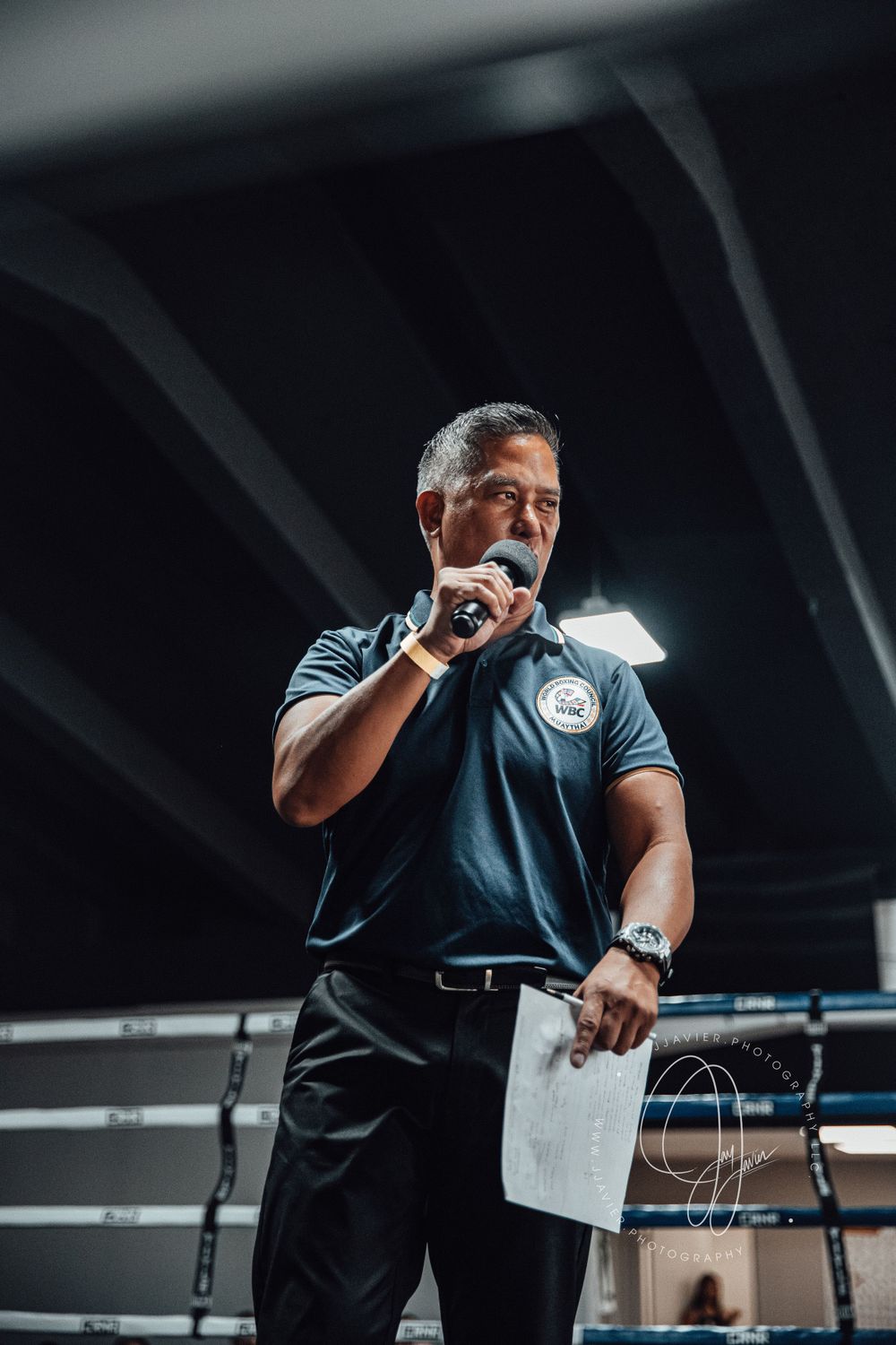 A referee in dark attire holds paperwork while officiating a sporting event in a dimly lit venue.