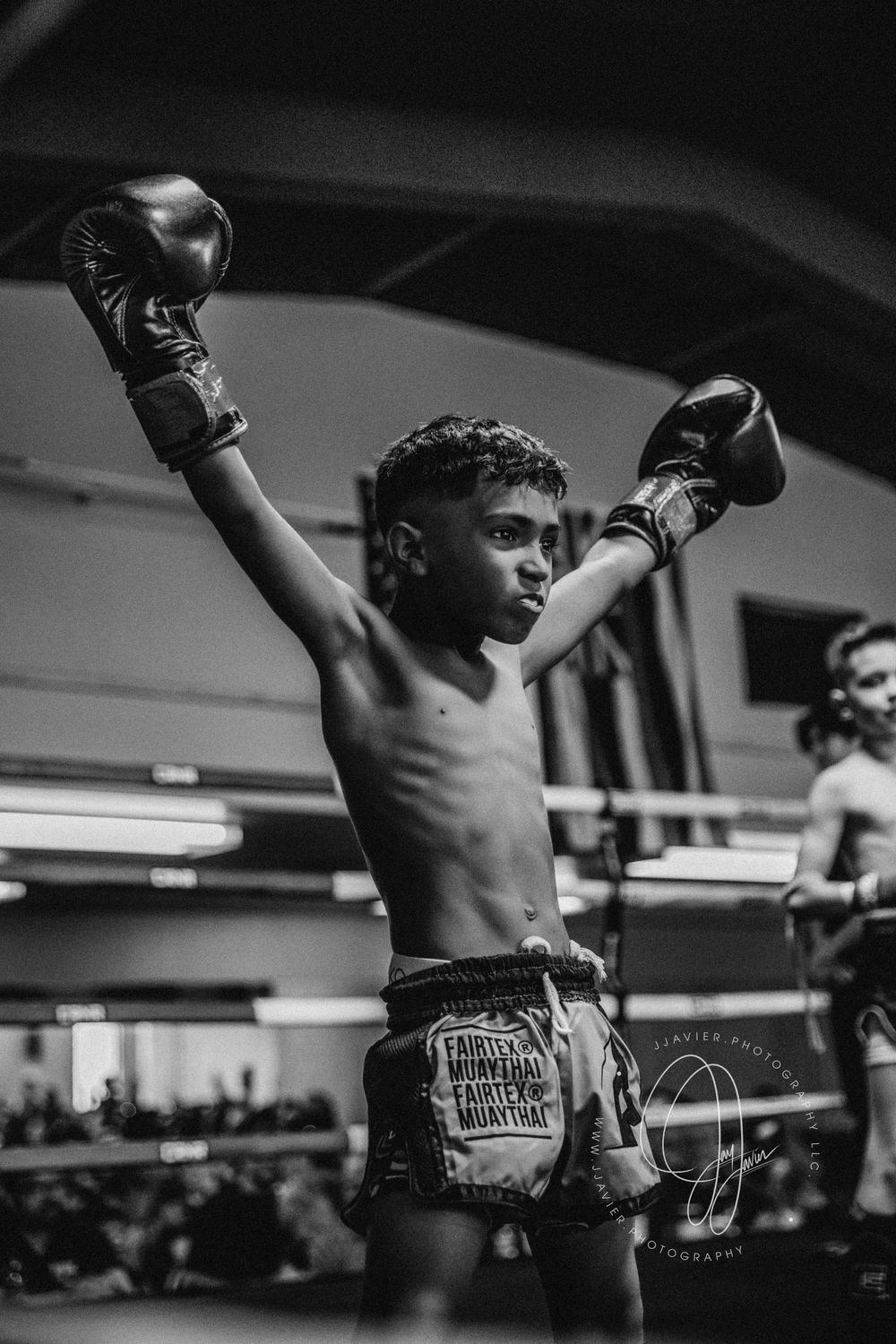 A young boxer raises their arms in victory wearing traditional Muay Thai boxing shorts.