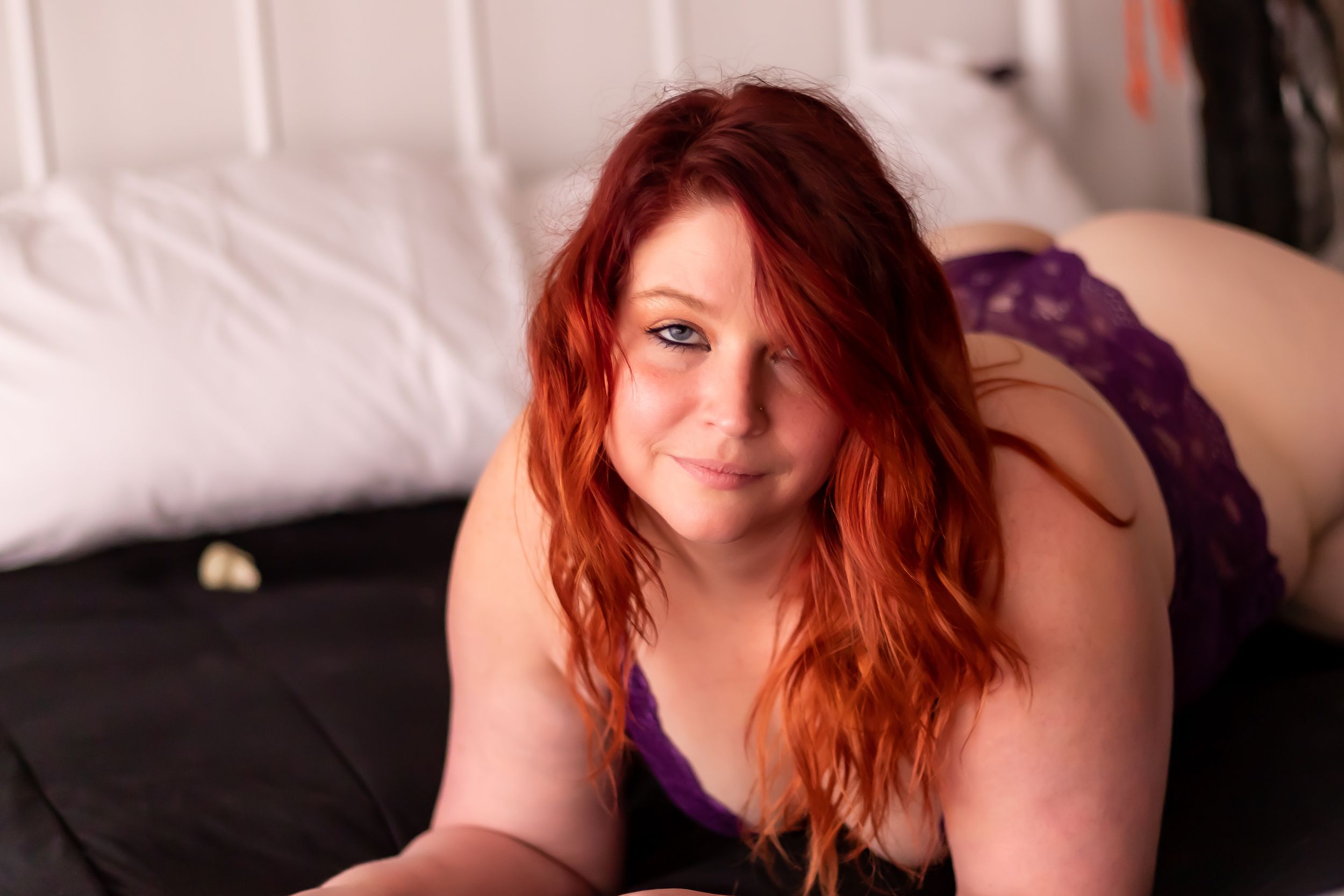 Intimate boudoir portrait with red hair and purple lingerie on dark bedding.