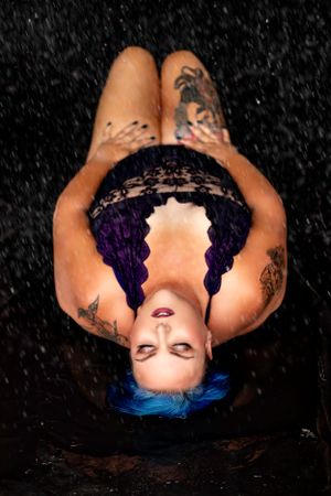 Creative portrait with water droplets against dark background featuring blue hair and tattoos.