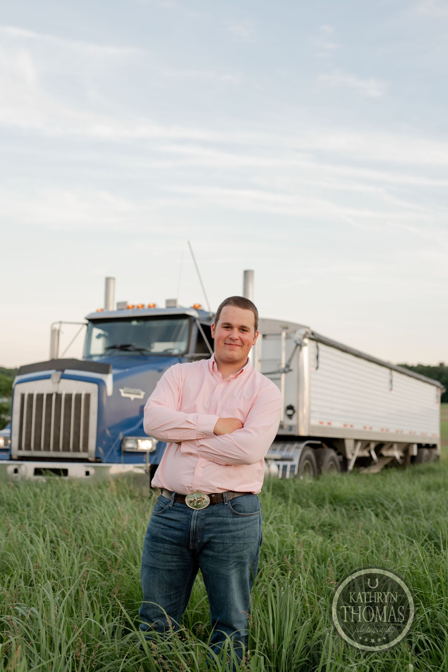 Professional portrait of a truck driver standing in front of a blue semi truck in a grassy field.