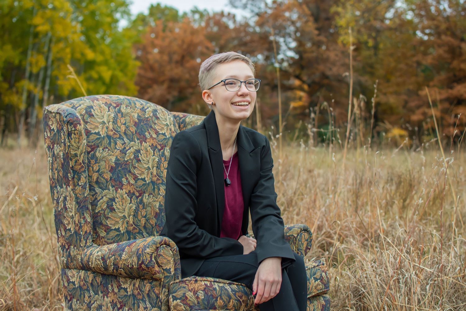 Person seated on floral patterned chair in autumn field wearing black jacket and glasses.