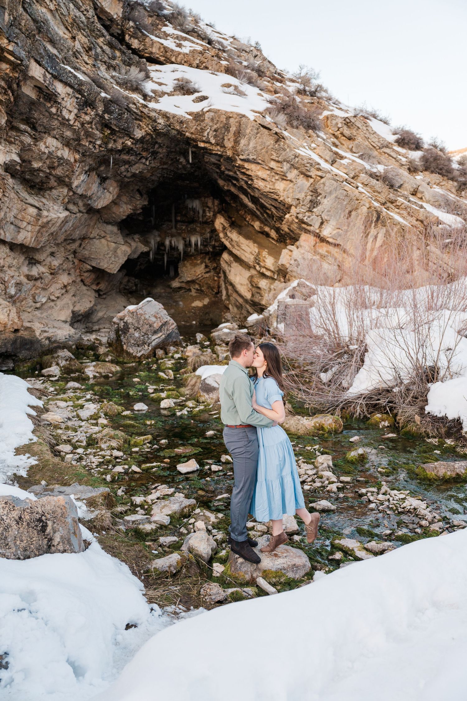 A romantic winter scene with a couple standing near a cave opening surrounded by snow and rocks.