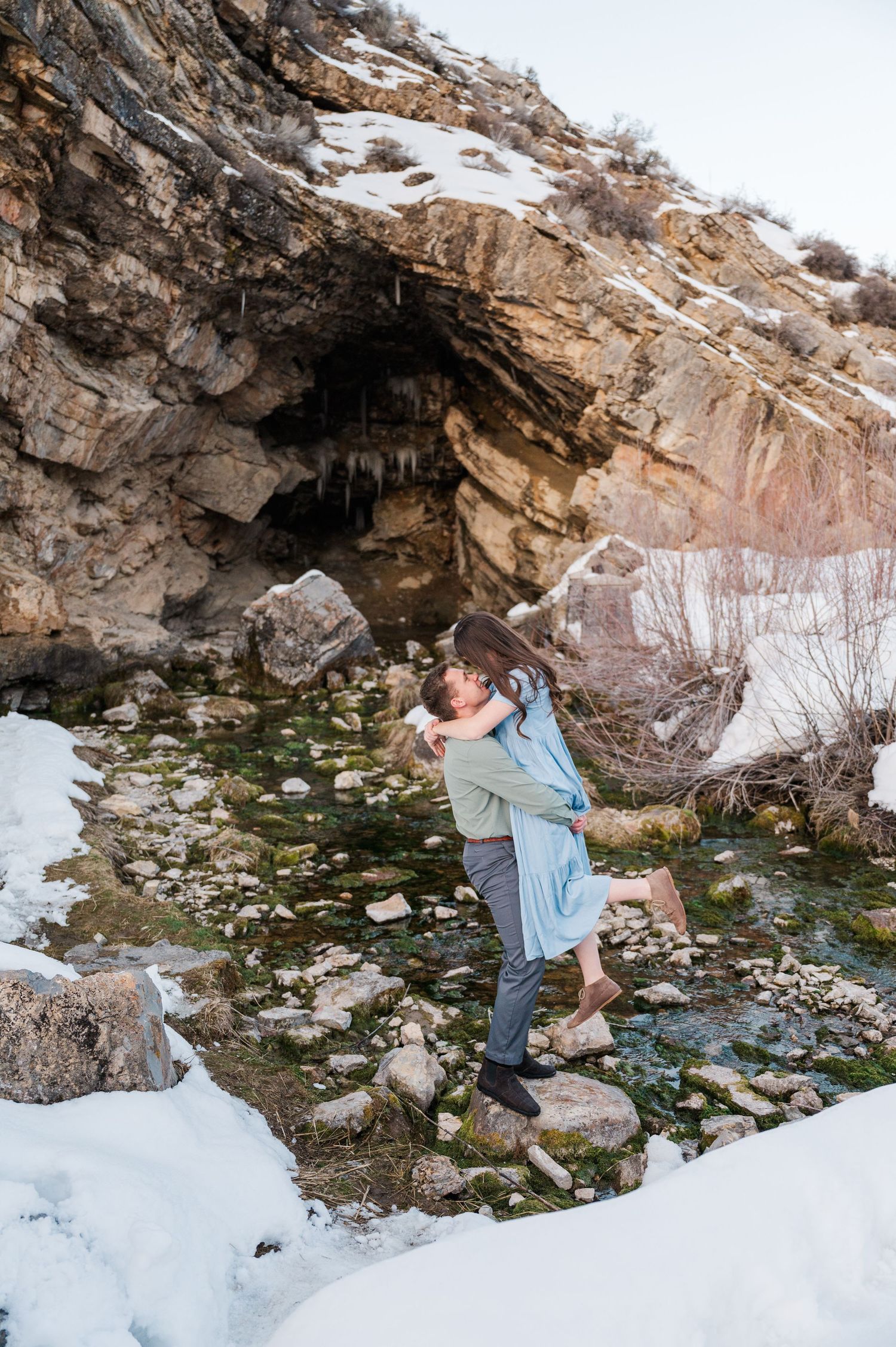 A romantic moment captured near a snow-covered cave entrance with rocky cliffs and scattered boulders.