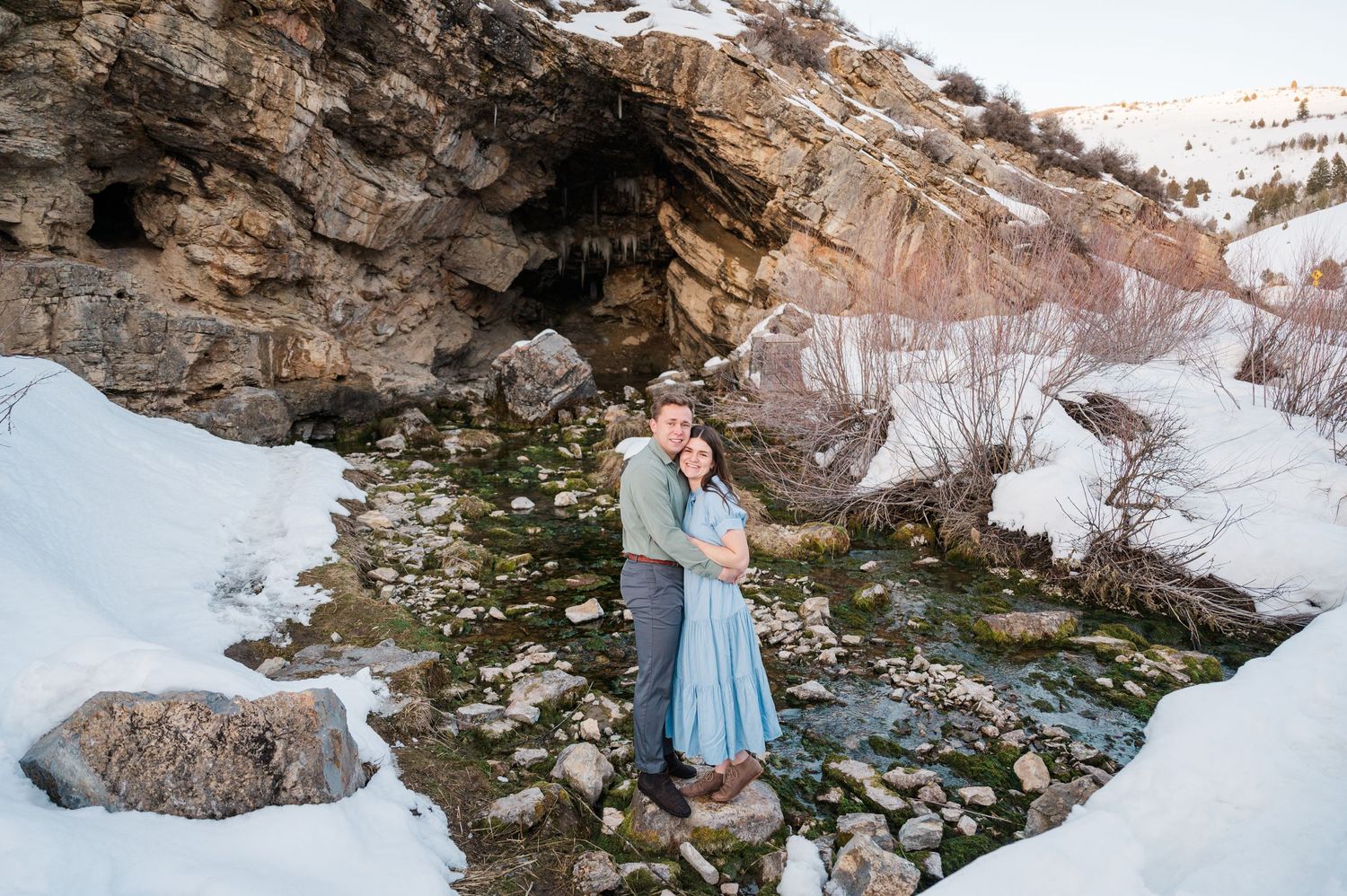 A couple embraces by a rocky cave entrance surrounded by snow and scattered stones.