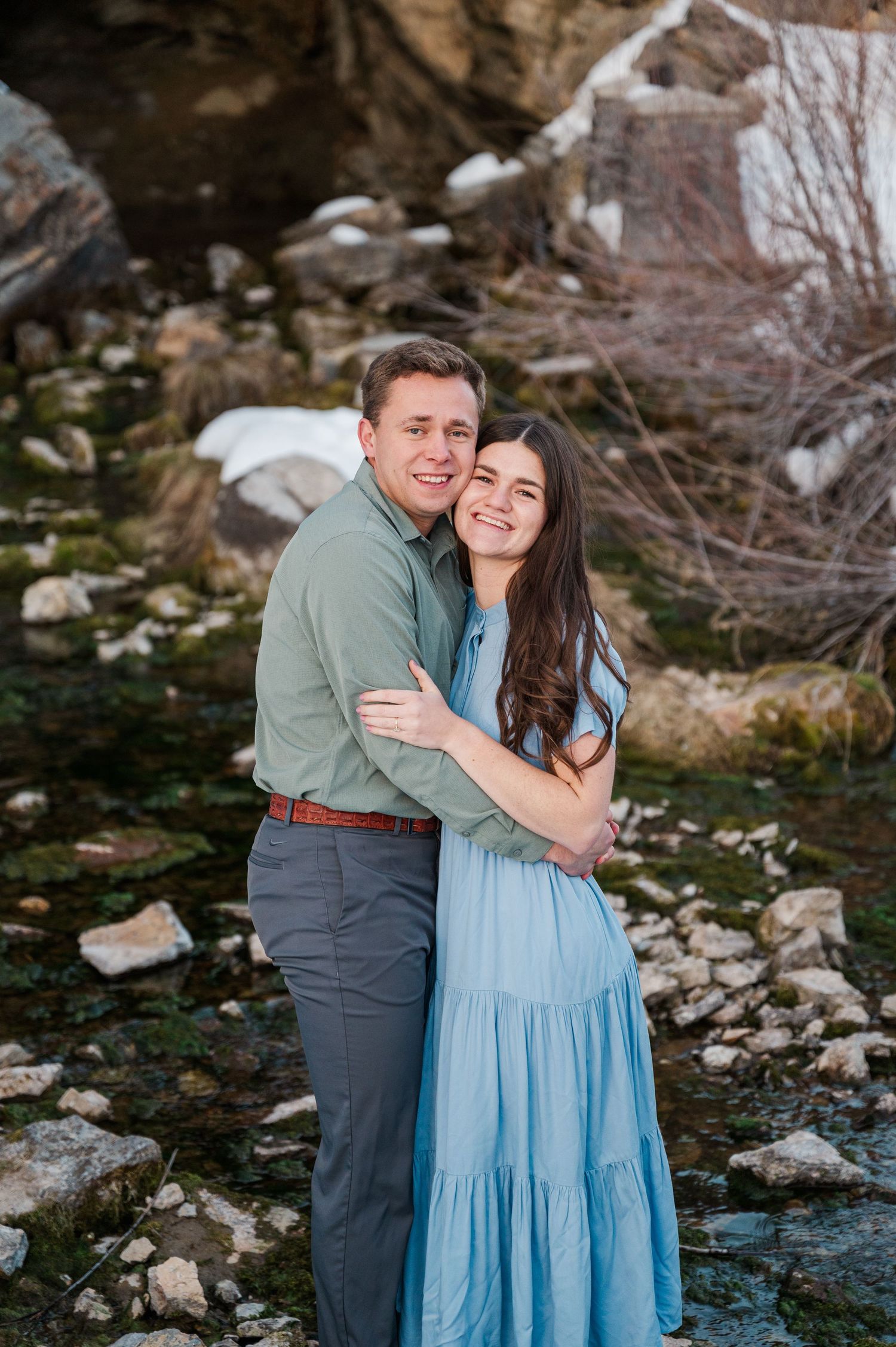 A couple in coordinating blue and sage outfits embraces near a rocky cave entrance.