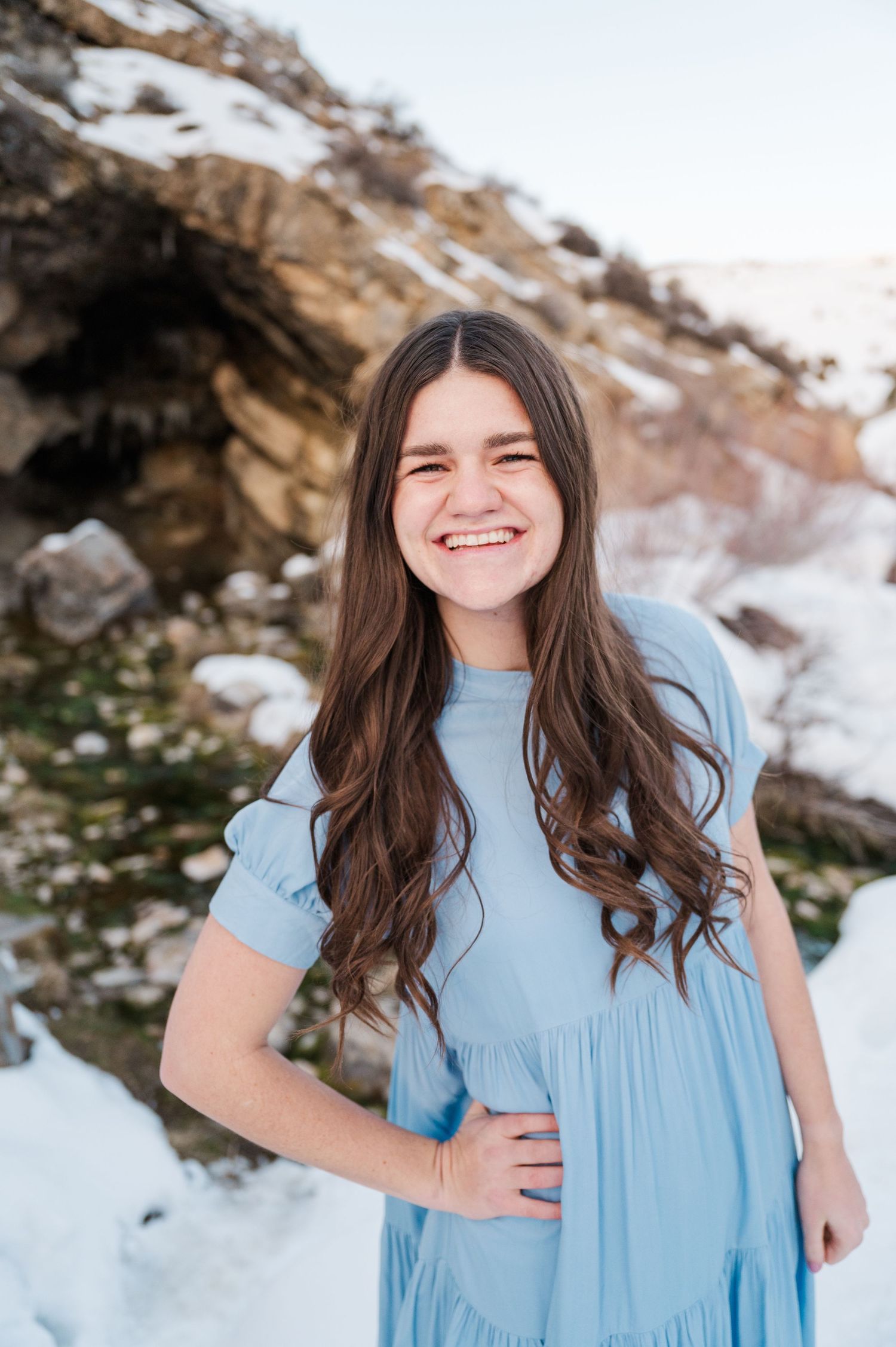 A woman in a light blue dress smiles against a snowy mountain backdrop.