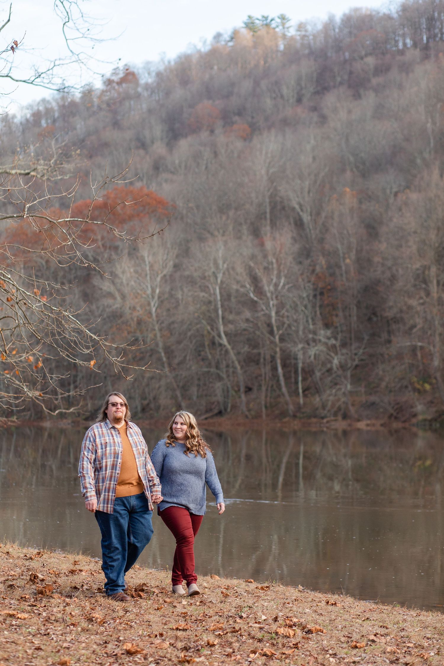 A couple shares a romantic moment by a serene lake surrounded by autumn trees and fallen leaves.