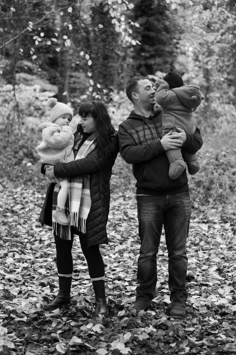A black and white image of a young family. Each parent is holding a child and they are looking at them, smiling.