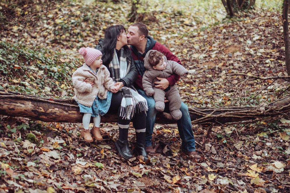 A young family sat on a fallen tree in the woods. The parents are having a kiss. In the brown autumn leaves.
