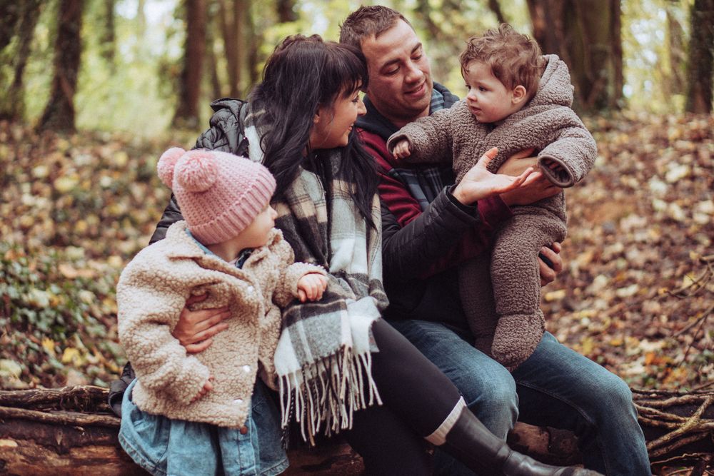 A young family are sat together on a fallen tree in woodland. They are all looking at the baby on dads lap.