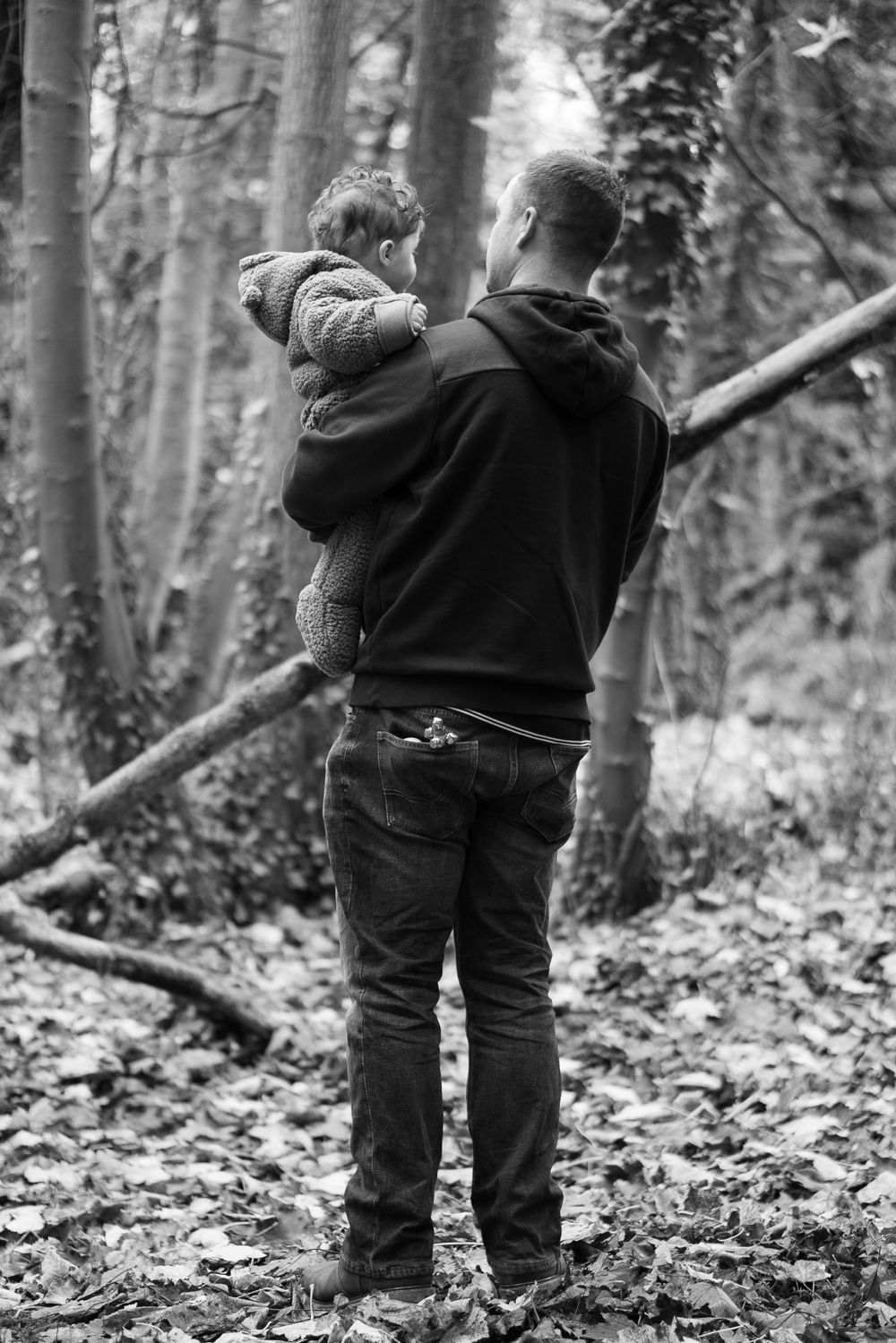 A black and white image. The back of a dad holding his baby in the woods.
