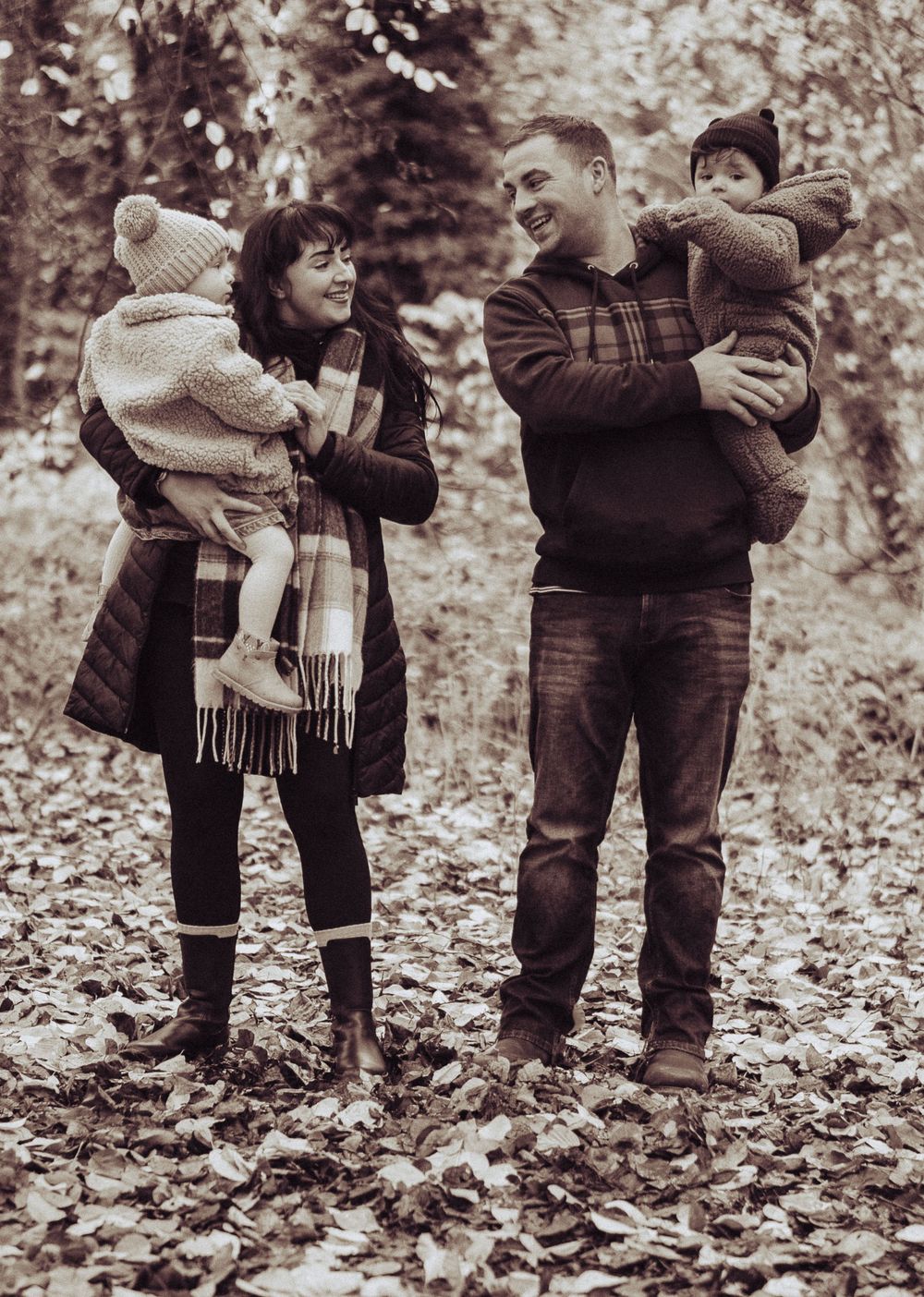 A black and white image of a young family. Each parent is holding a child, the parents are looking at each other.