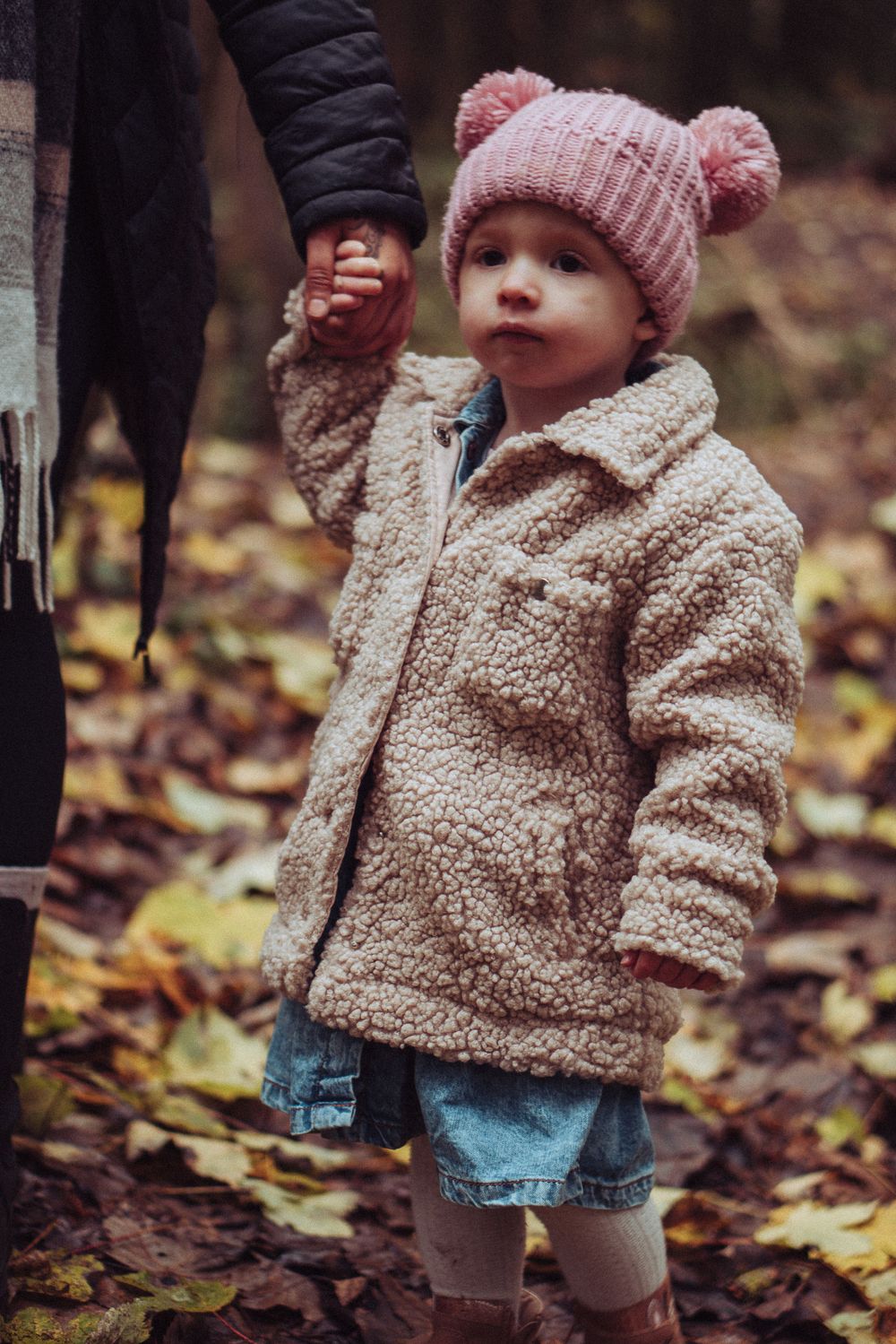 A little girl looking into the distance, holding her mums hand. She is wrapped up in a fluffy coat and a pink hat.