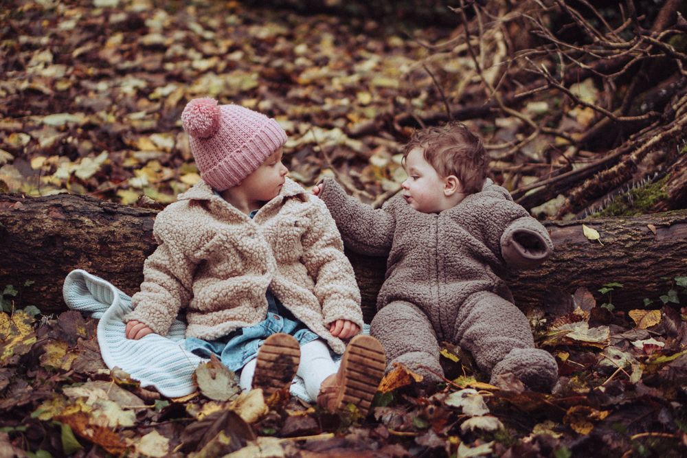 A young girl and her baby brother sat in the autumn leaves, in front of a fallen tree in the woods.
