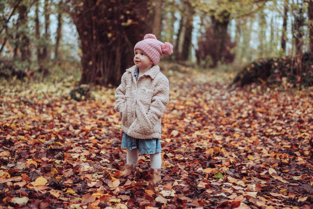 A little girl stood in the autumn leaves, with her hands in her pockets of her coat.