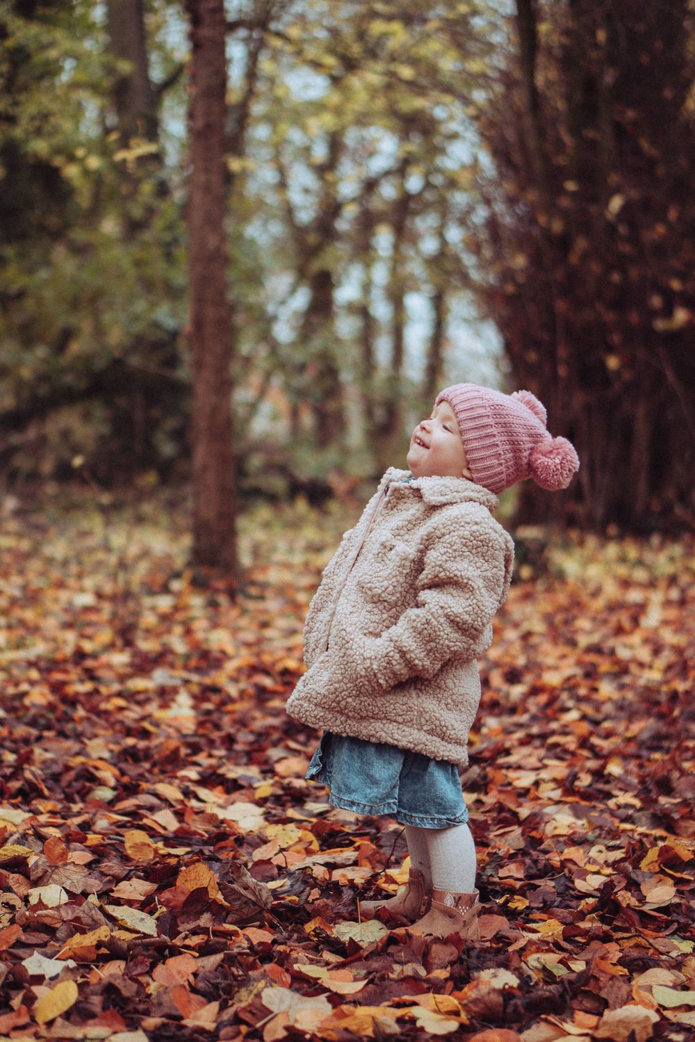 A little girl standing with her hands in her pockets, leaning back and laughing. In the autumn leaves.