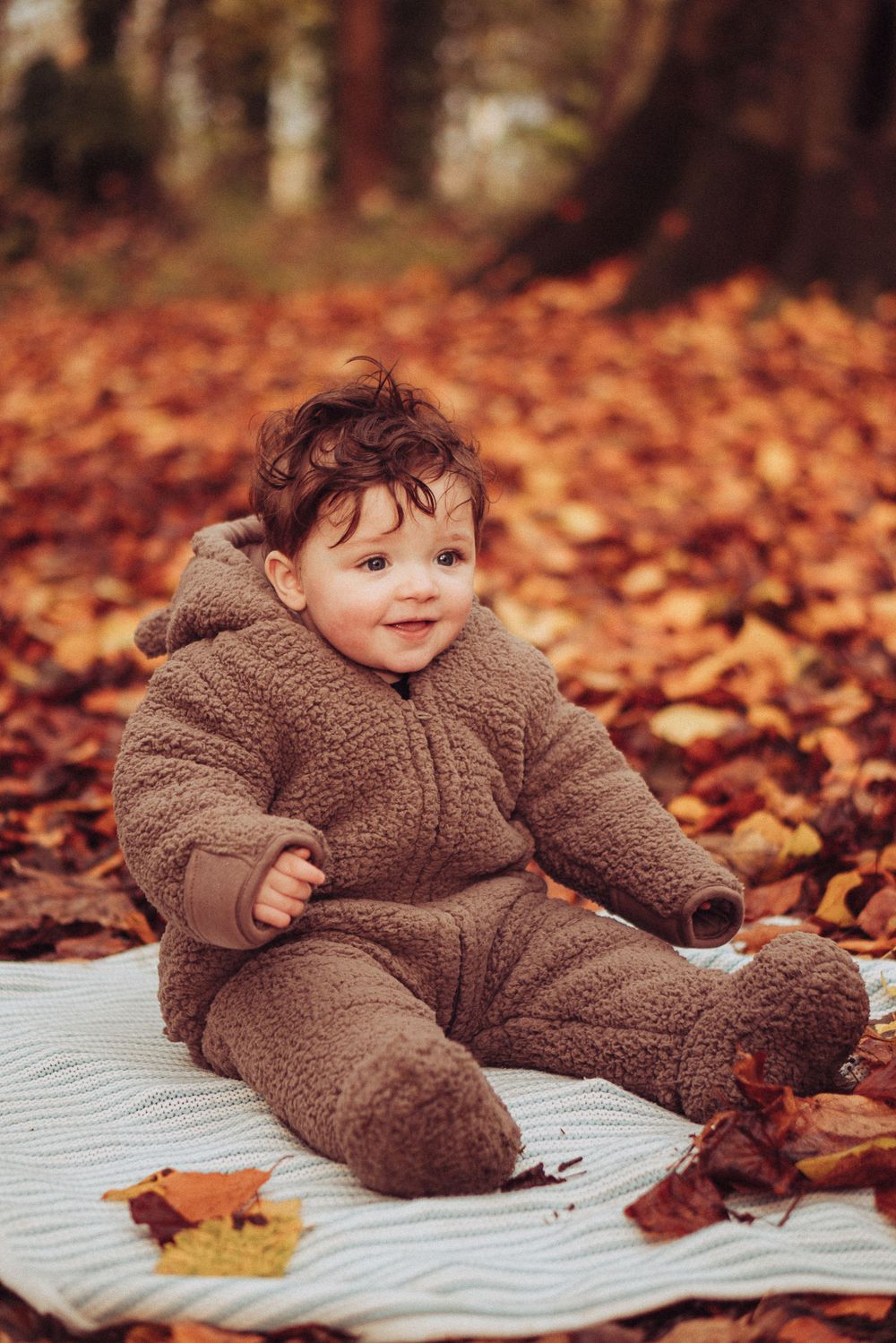 A little boy wrapped up in a bear onesie, sat on a blanket in the autumn leaves. He has the biggest smile.