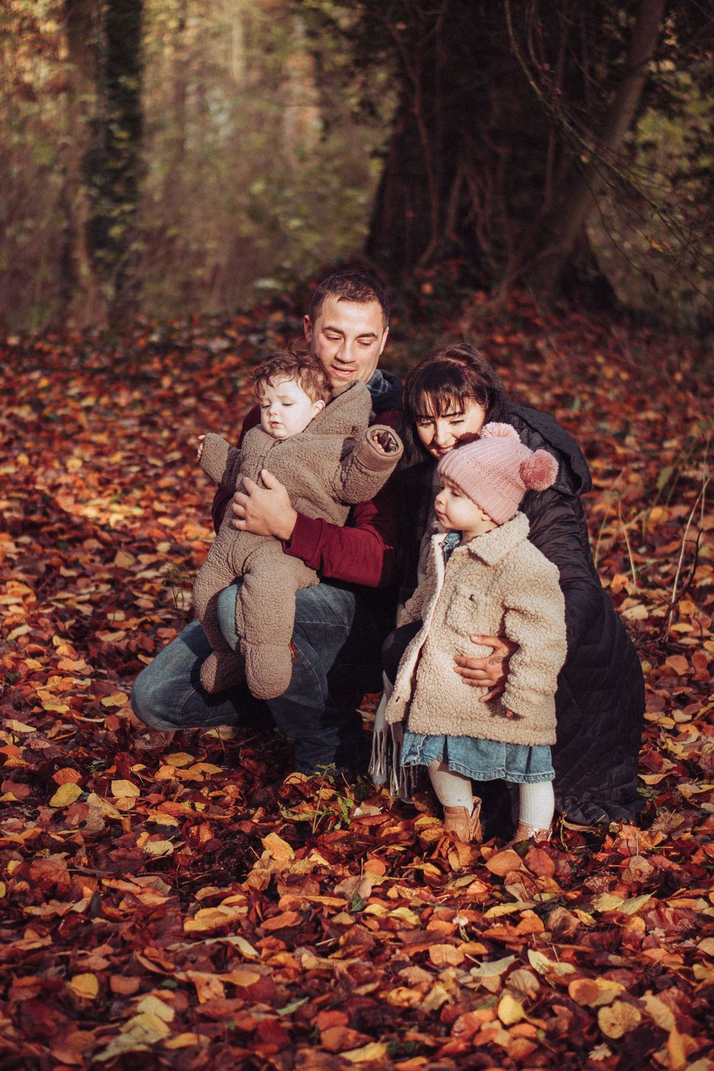 A young family are crouched down together in the brown autumn leaves.