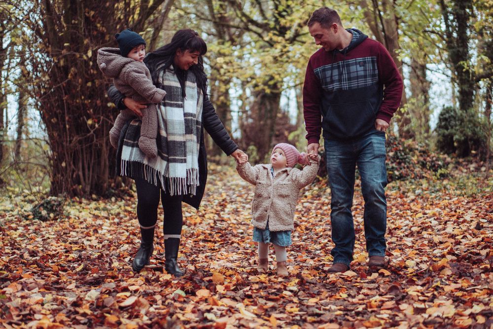A young family walking towards the camera, through the brown autumn leaves, all holding hands. The parents are both looking down.