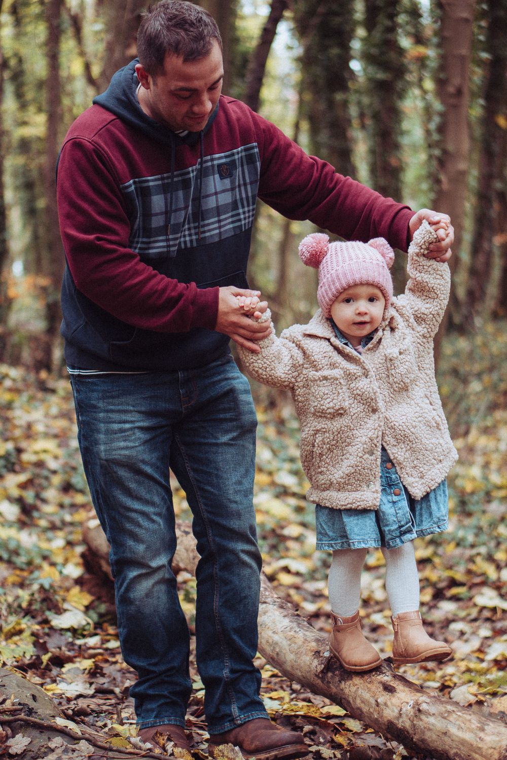 A dad holding his little girls hands as she walks along a fallen tree in the woods.
