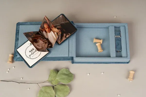 Light blue photo box with scattered prints and green leaves on a neutral surface.