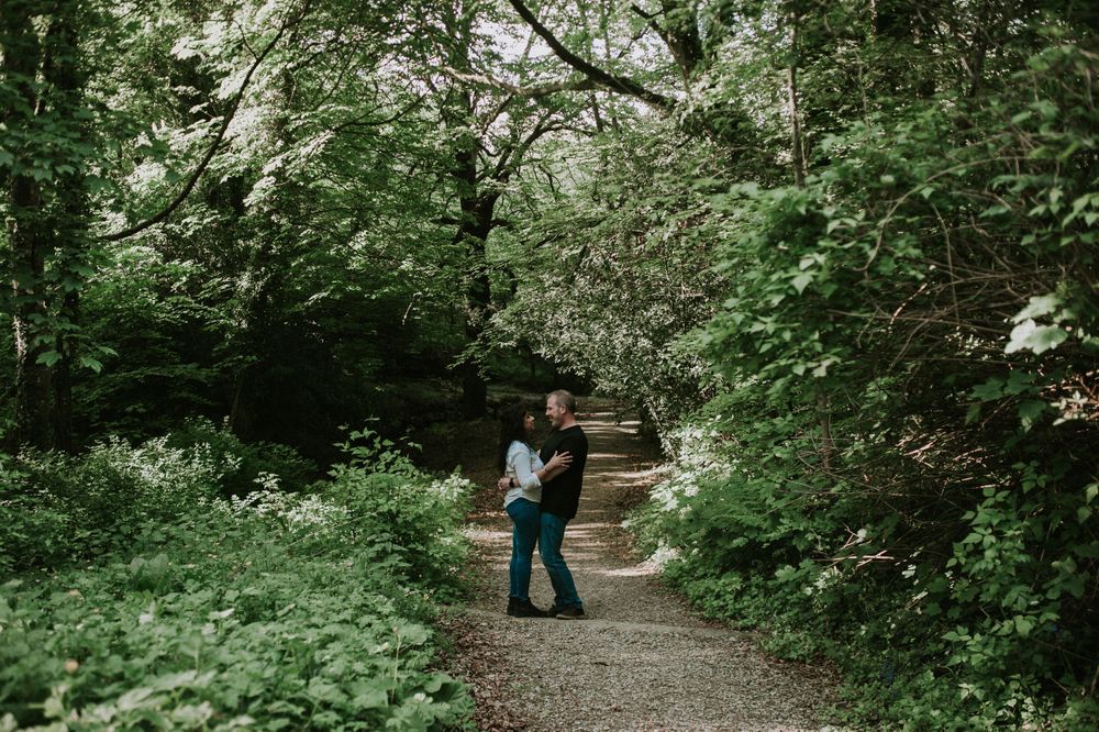 A couple embracing on a tree-lined walking path in a lush green forest.